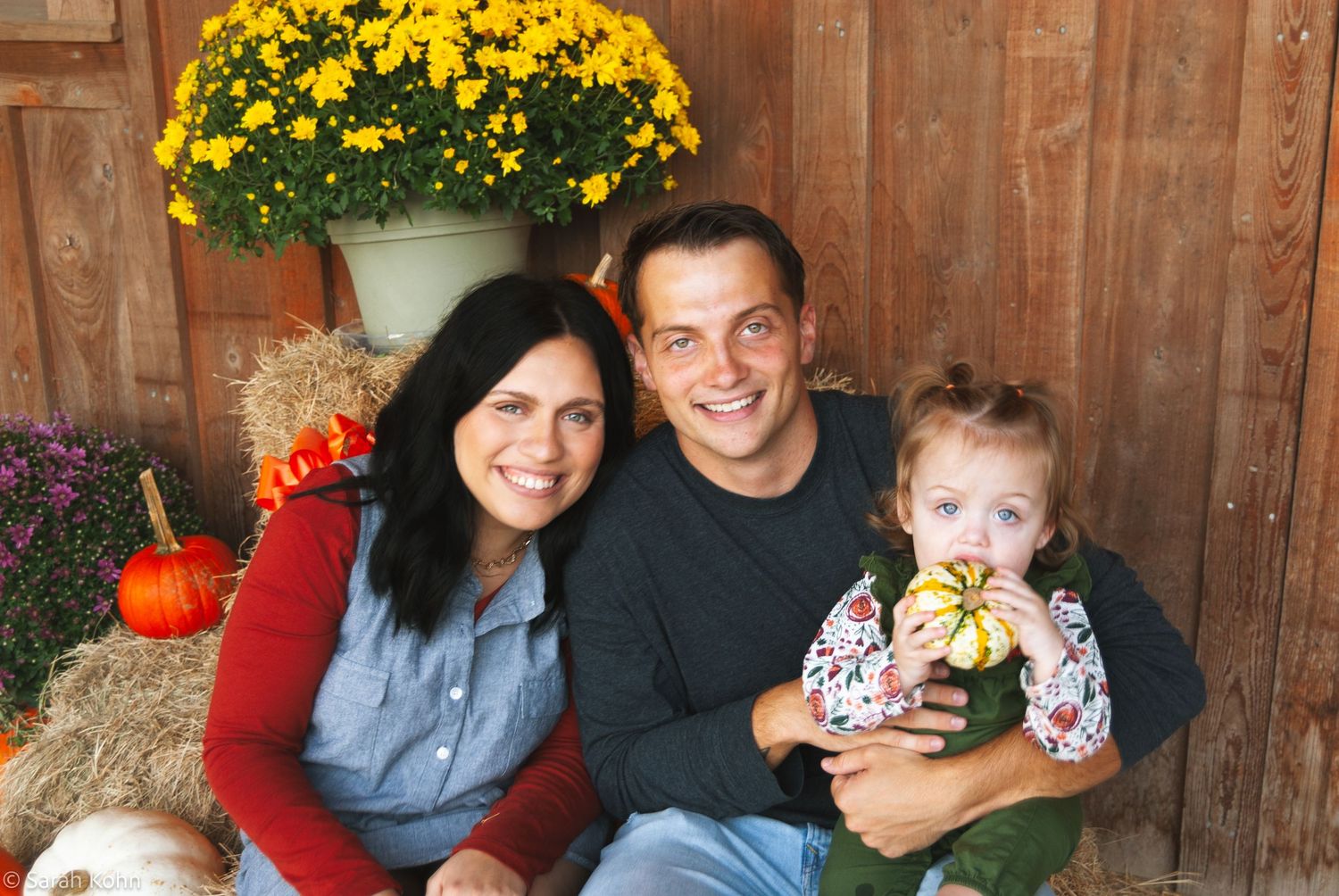 Happy family with baby posing among fall pumpkins and yellow mums for autumn portrait.