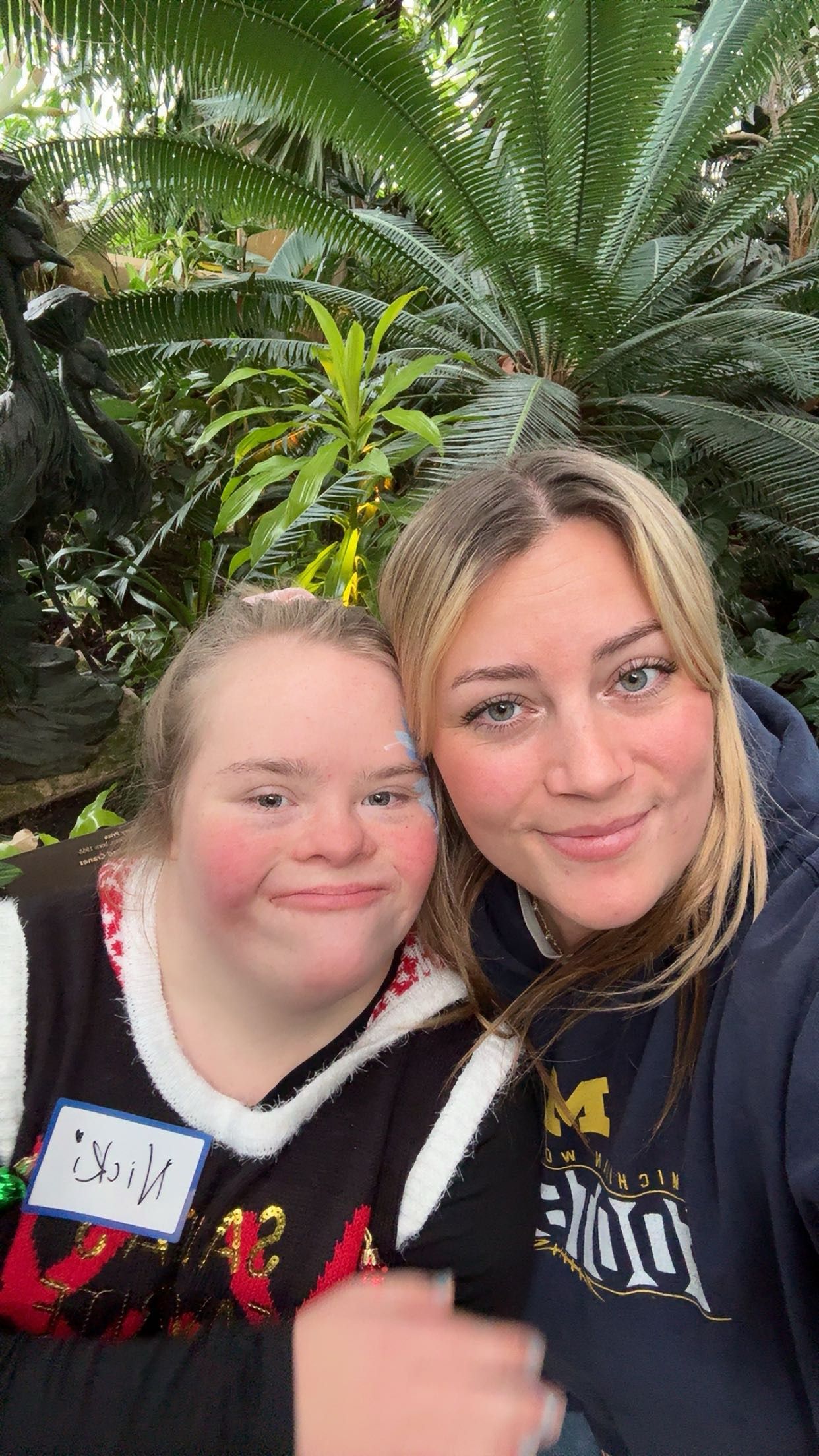 Two young women smiling together in a lush tropical greenhouse, one wearing a name tag and festive sweater.