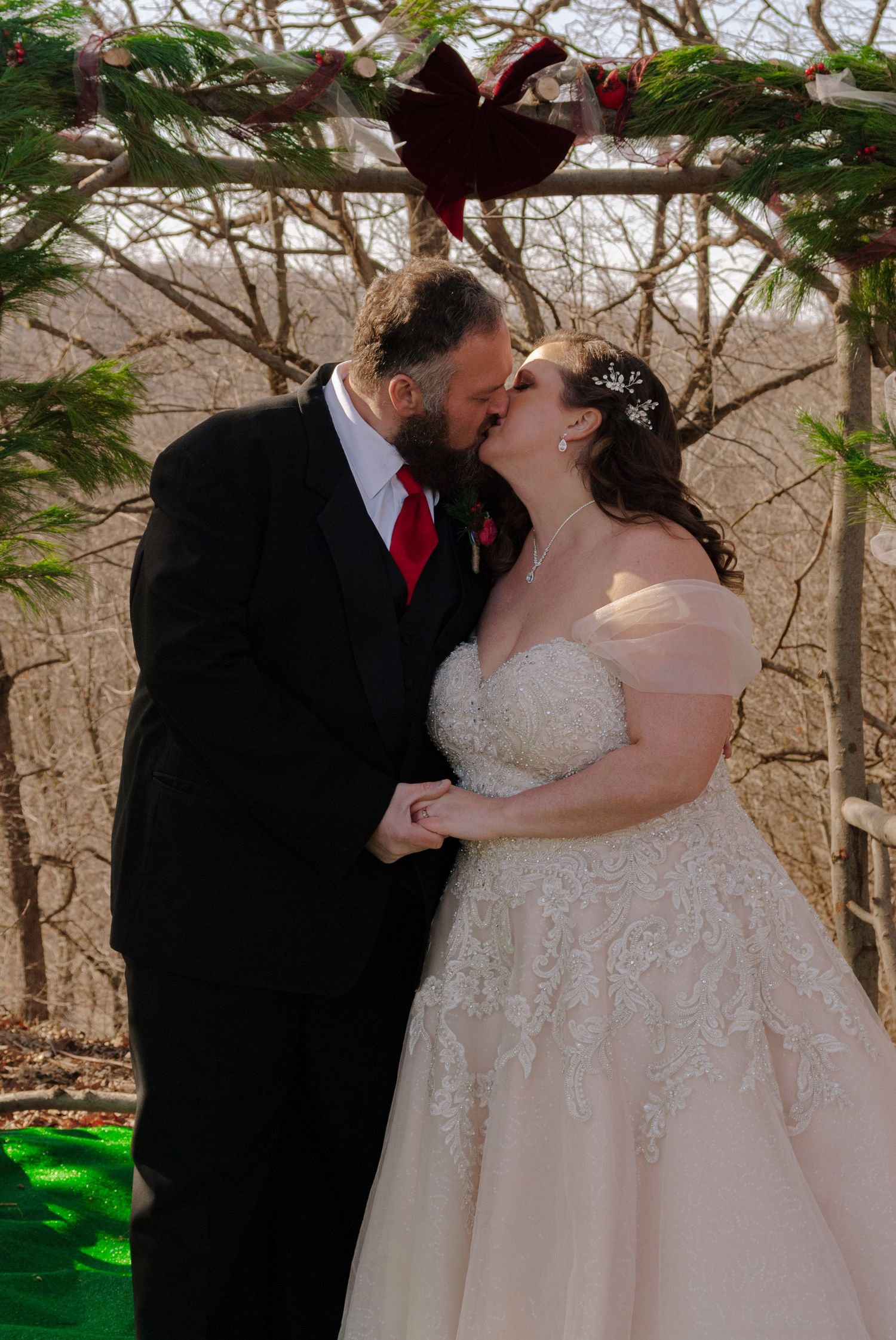 Bride and groom sharing first kiss at outdoor winter wedding ceremony under pine arch.