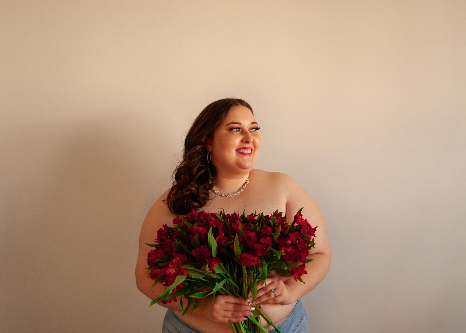 Smiling plus-size woman holds a large bouquet of red alstroemeria flowers against a neutral wall.