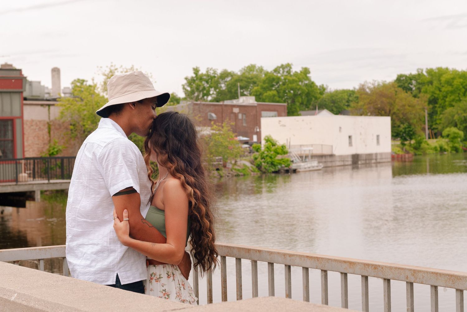 Loving couple kissing on bridge overlooking flooded river with green trees and buildings in background.