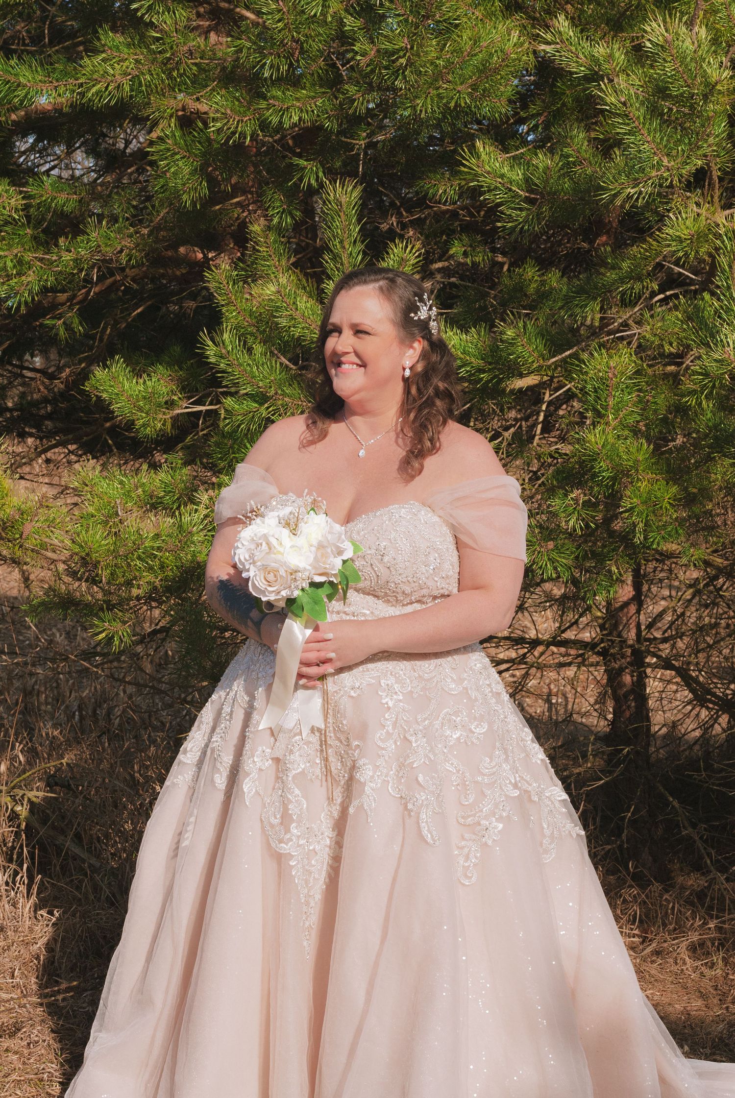 Bride in off-shoulder lace ball gown holds white bouquet beside evergreen tree.