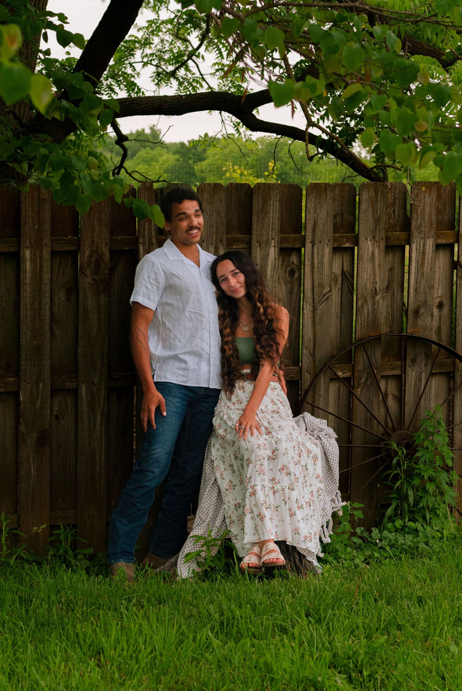 A smiling couple standing together under a tree branch near a rustic wooden fence and wagon wheel.