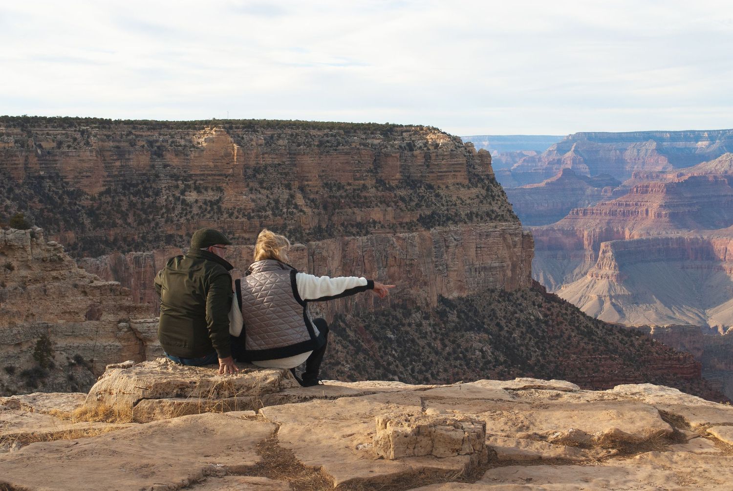 Couple sitting on rocky ledge overlooking the vast Grand Canyon landscape, one person pointing into the distance.