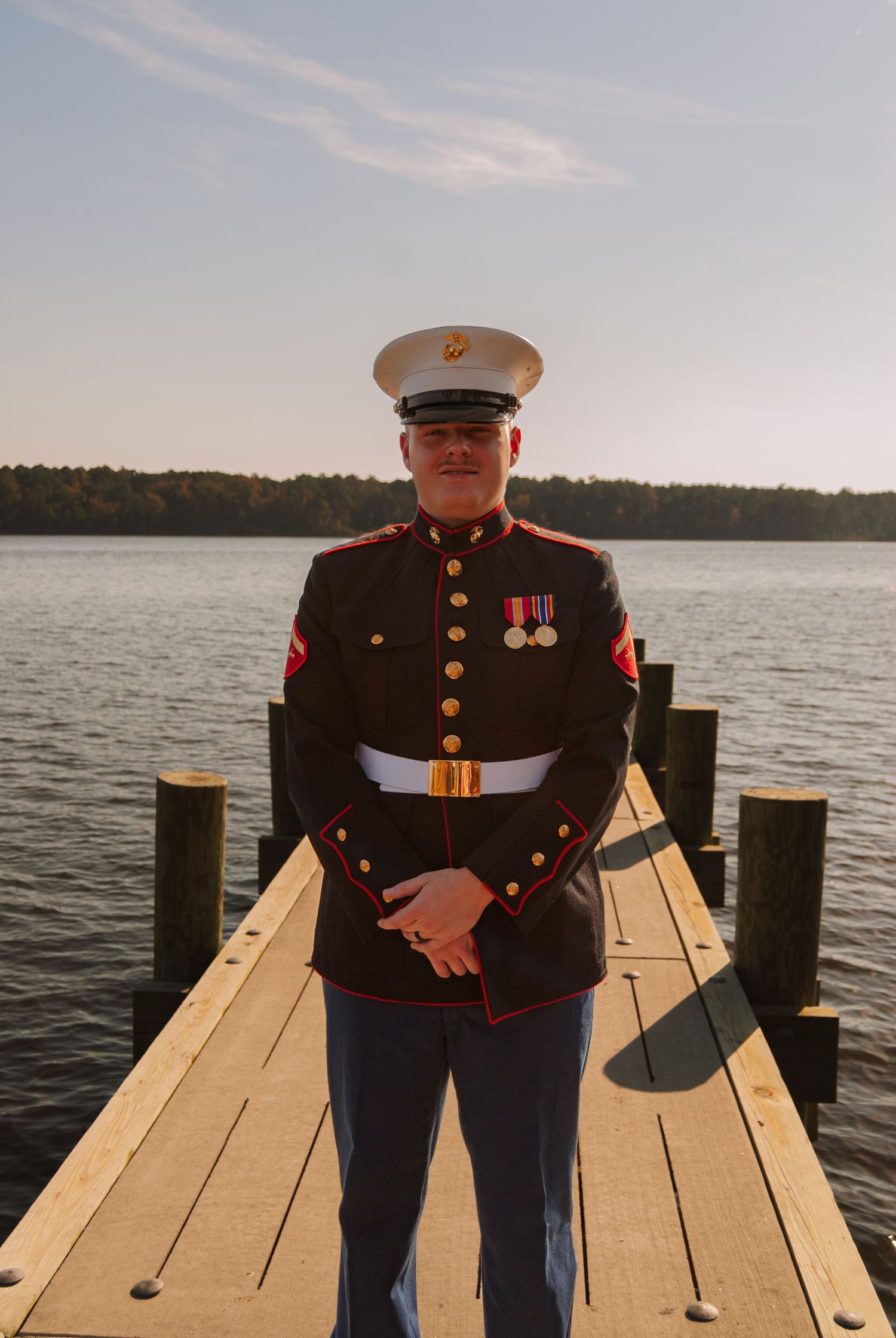 US Marine in dress blue uniform standing proudly on wooden dock with lake backdrop.