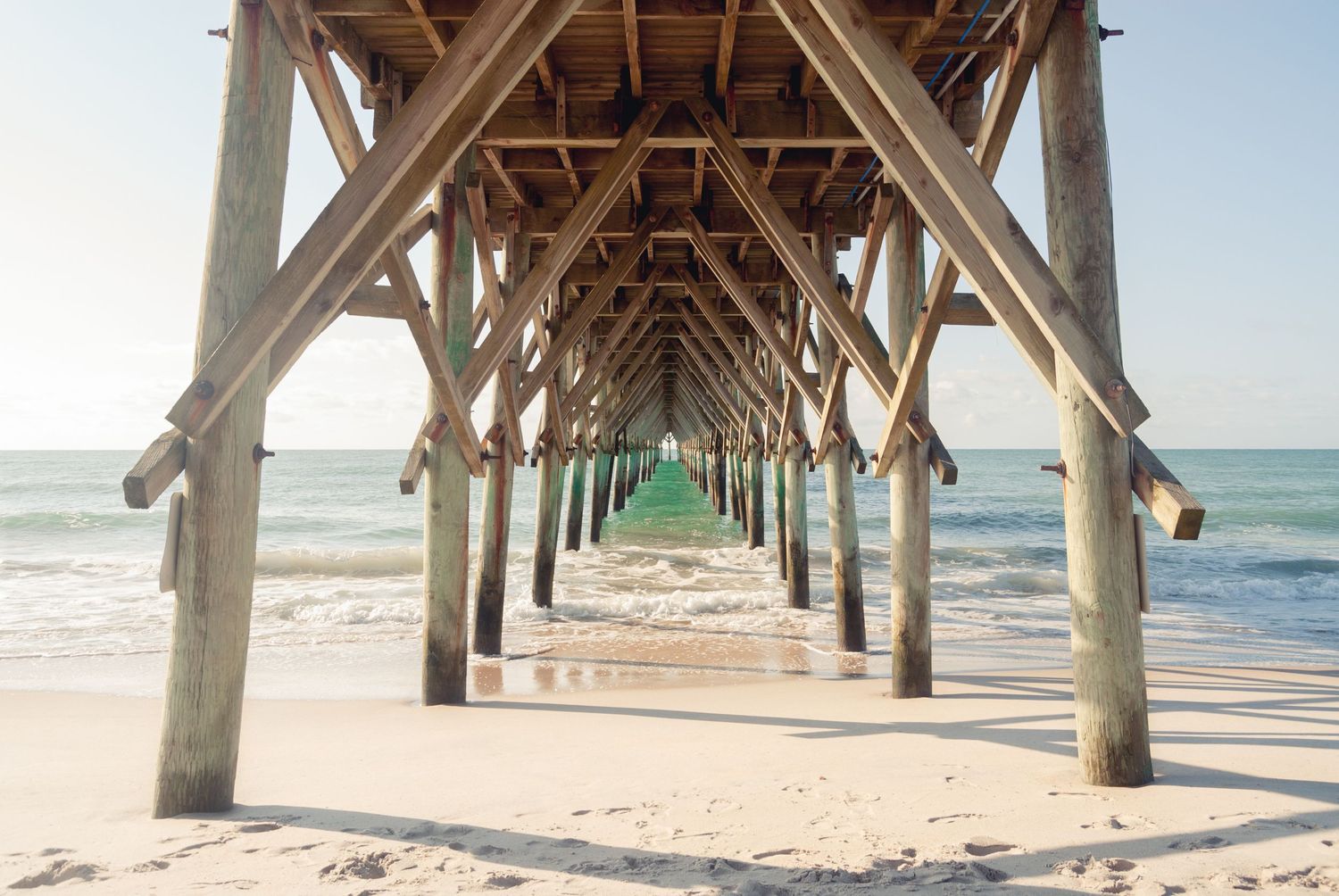Symmetrical view under wooden beach pier with sandy shore and turquoise ocean beyond.