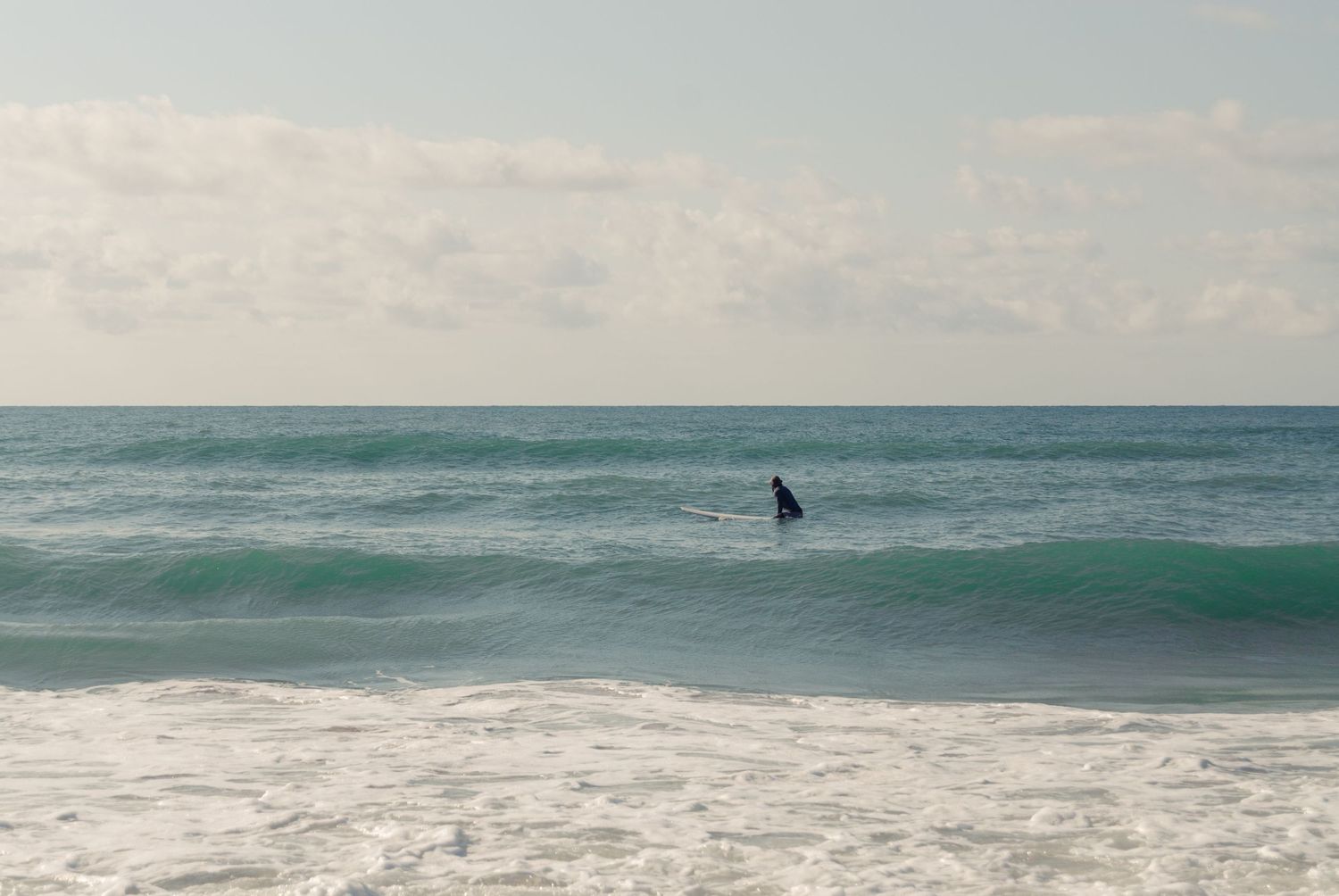 Lone surfer standing on board in calm turquoise ocean waters under cloudy sky.