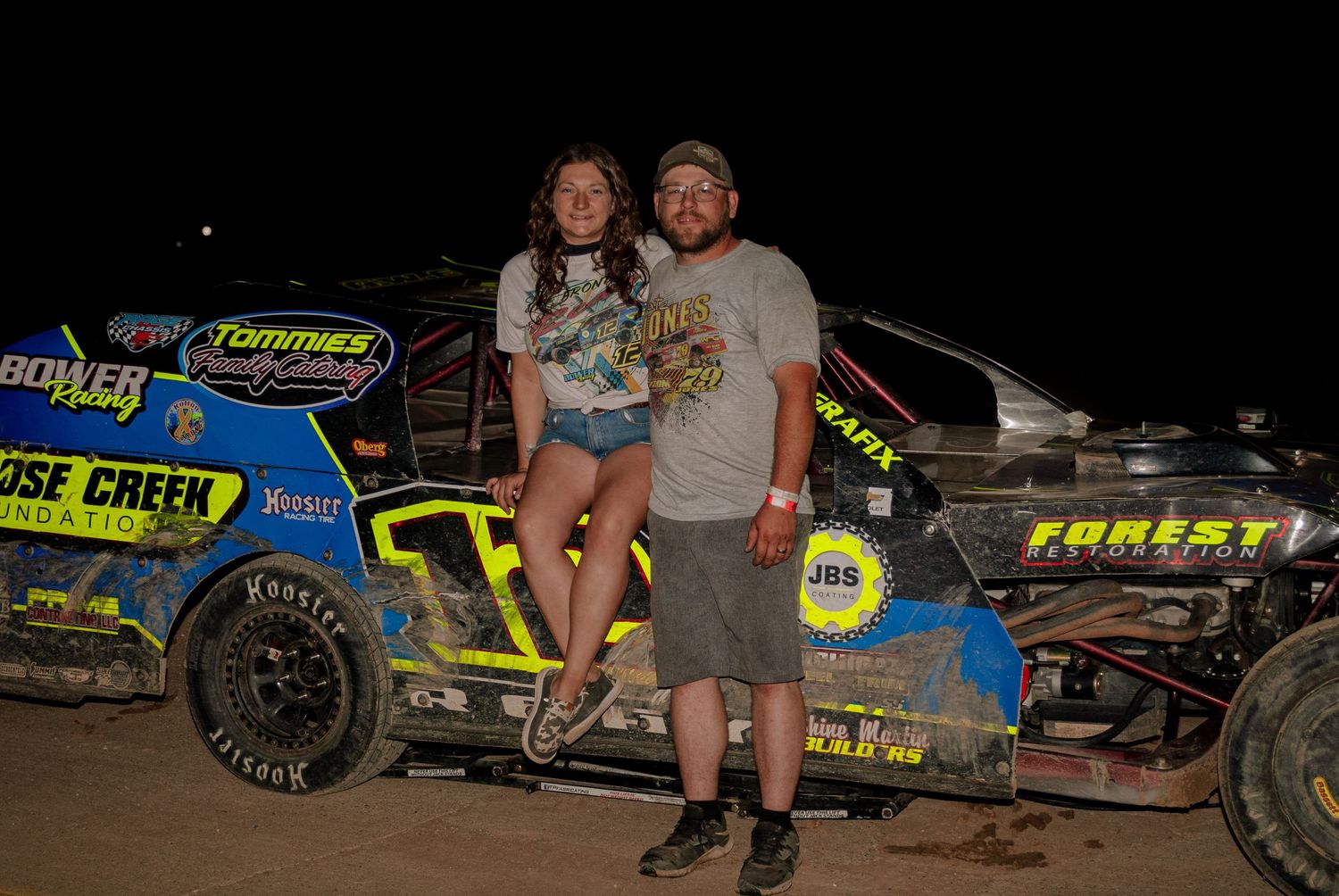 Smiling couple posing together in front of a sponsored dirt track modified race car at night.