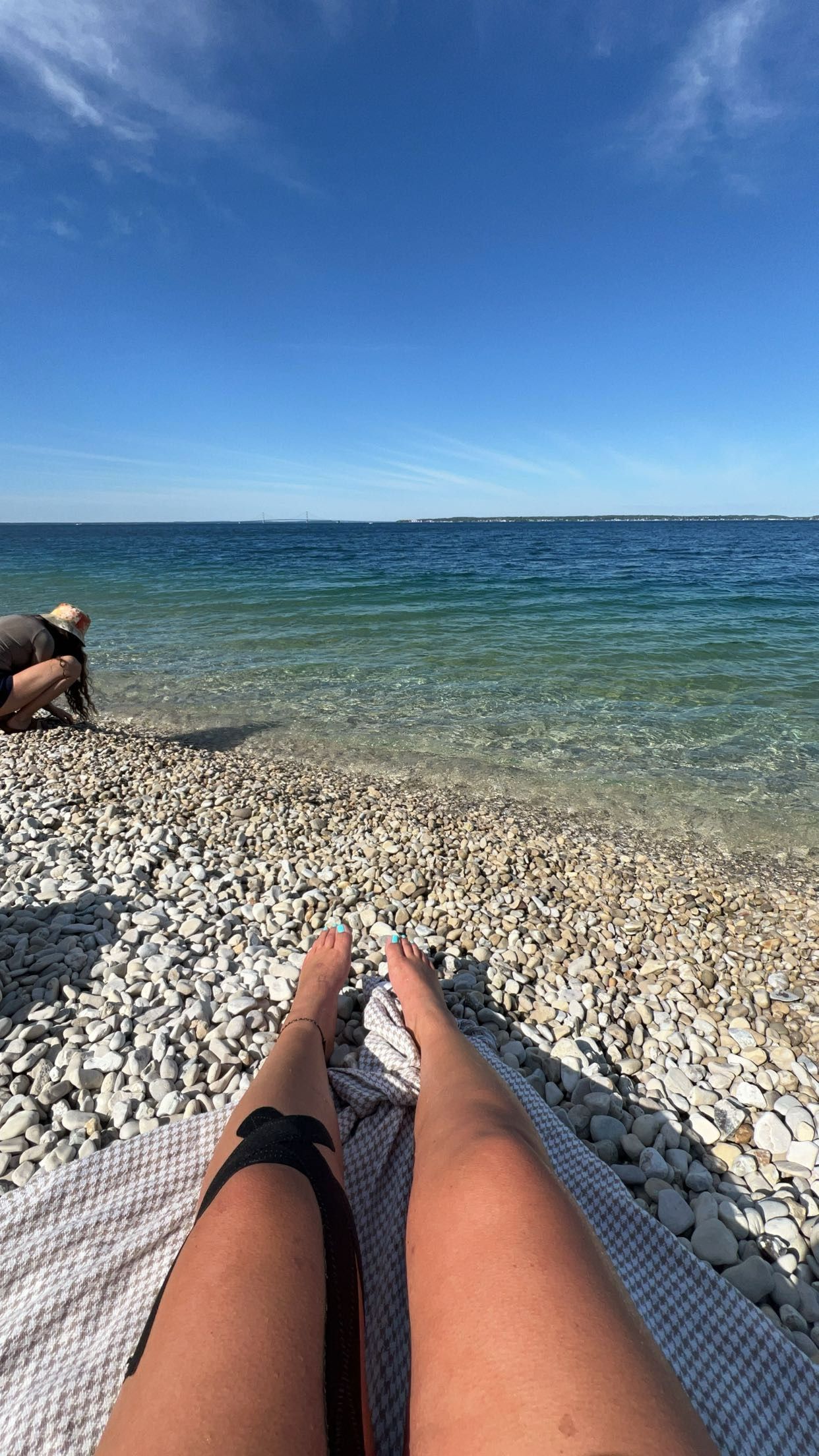Person relaxing on a rocky pebble beach with clear turquoise water and blue sky, legs stretched out on a checkered blanket.