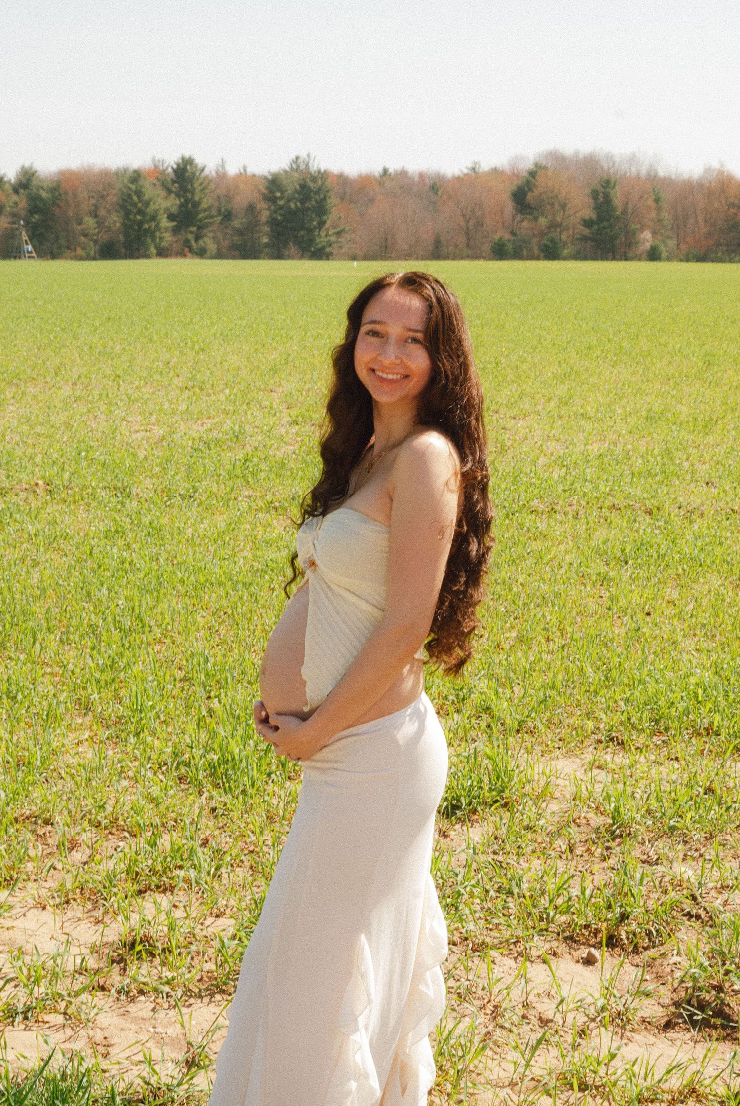 Pregnant woman in white dress holding baby bump while standing in green field.