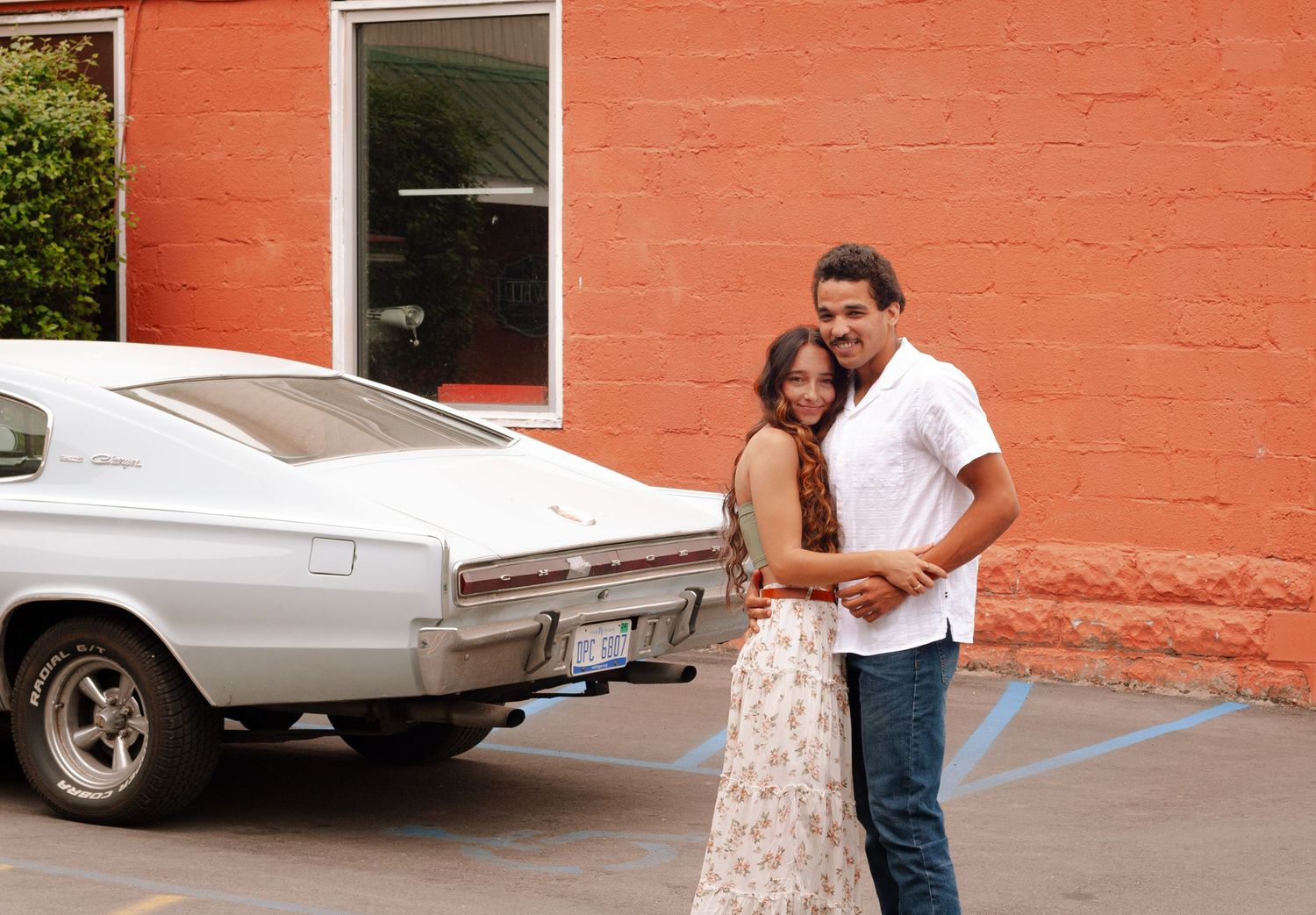 Happy couple embracing and smiling beside a vintage white car parked near an orange building.