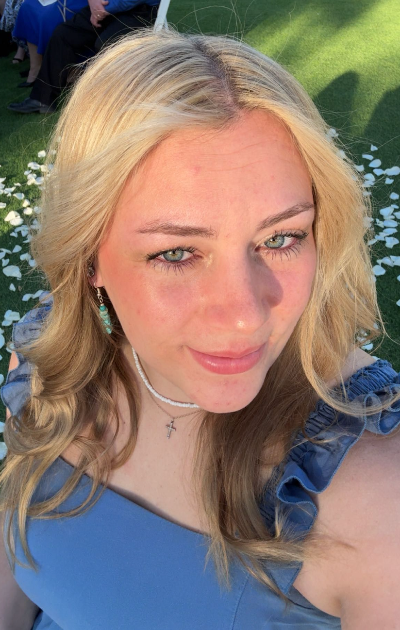 Young blonde woman with green eyes wearing a blue ruffled top and cross necklace takes a sunny outdoor selfie.
