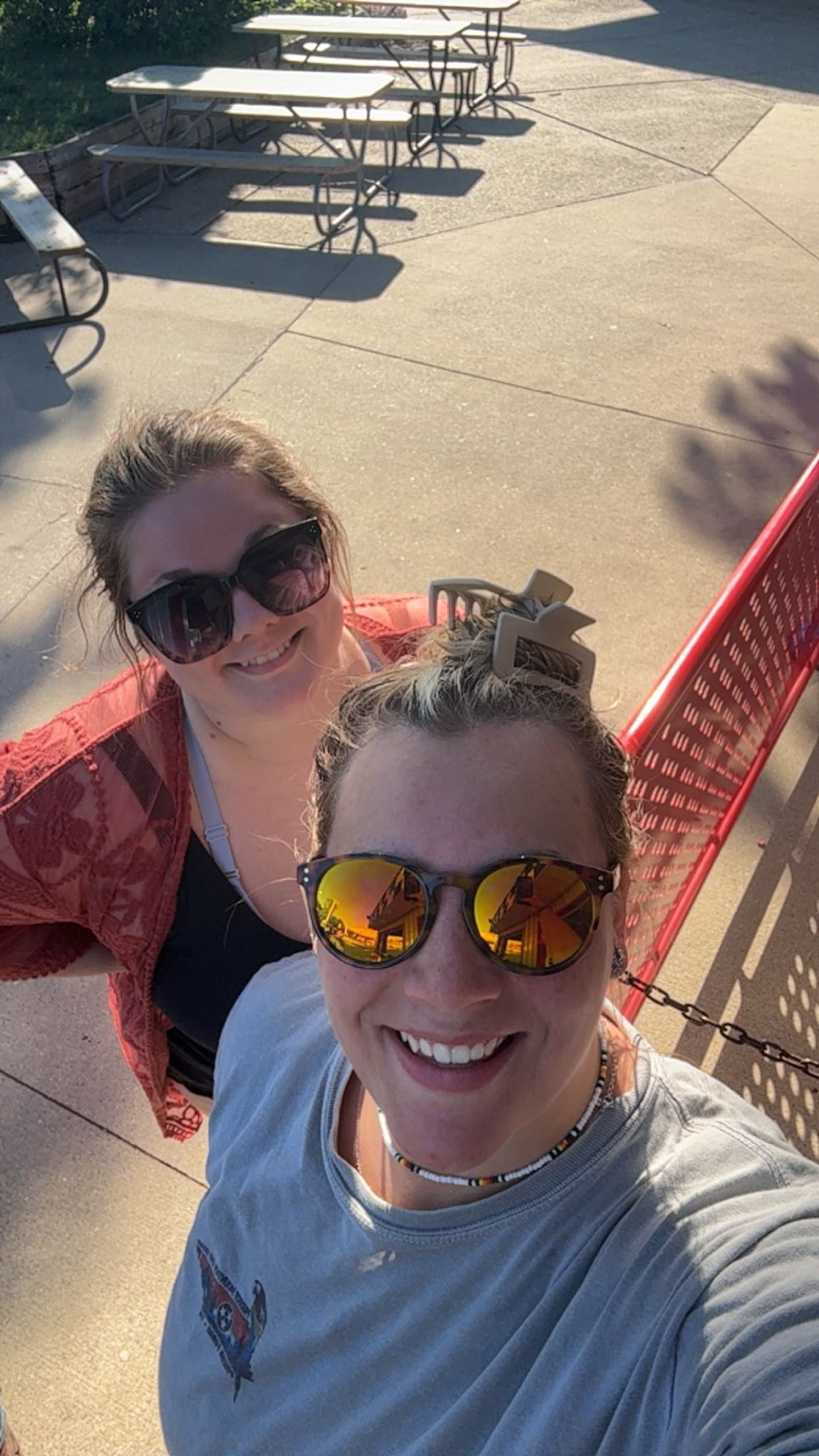 Two smiling women taking a selfie outdoors at a patio area with red chairs and picnic tables in the background.
