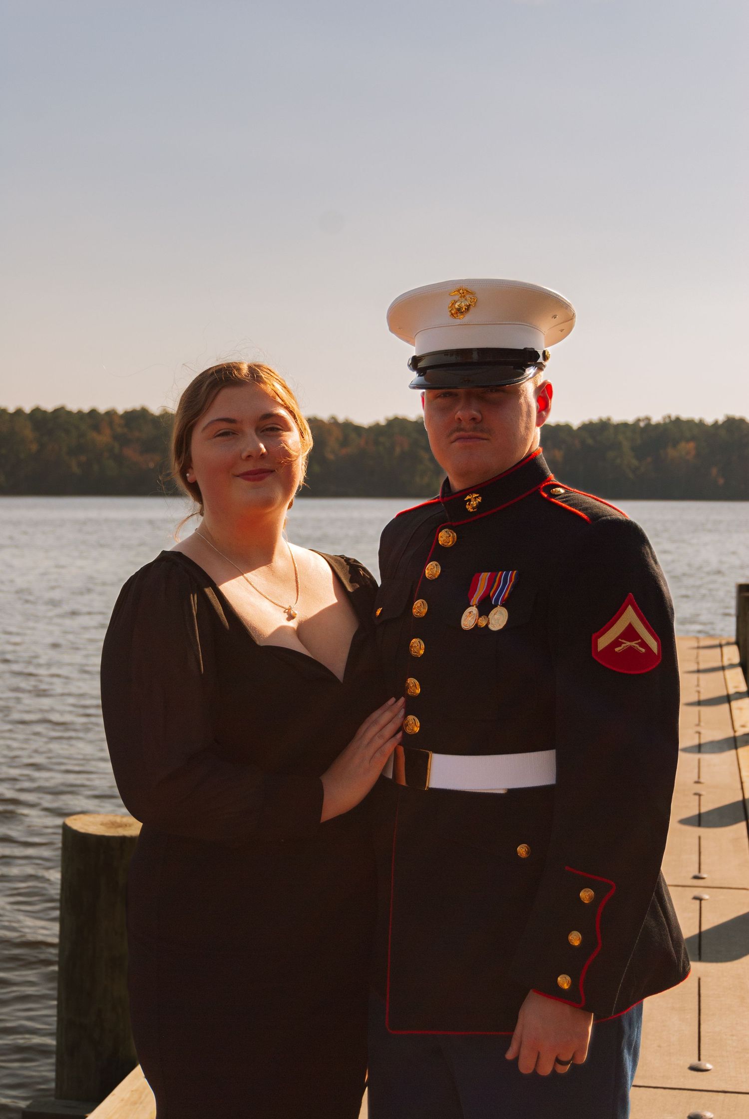 A woman in black dress poses with a US Marine in dress blues uniform on a lakeside dock.