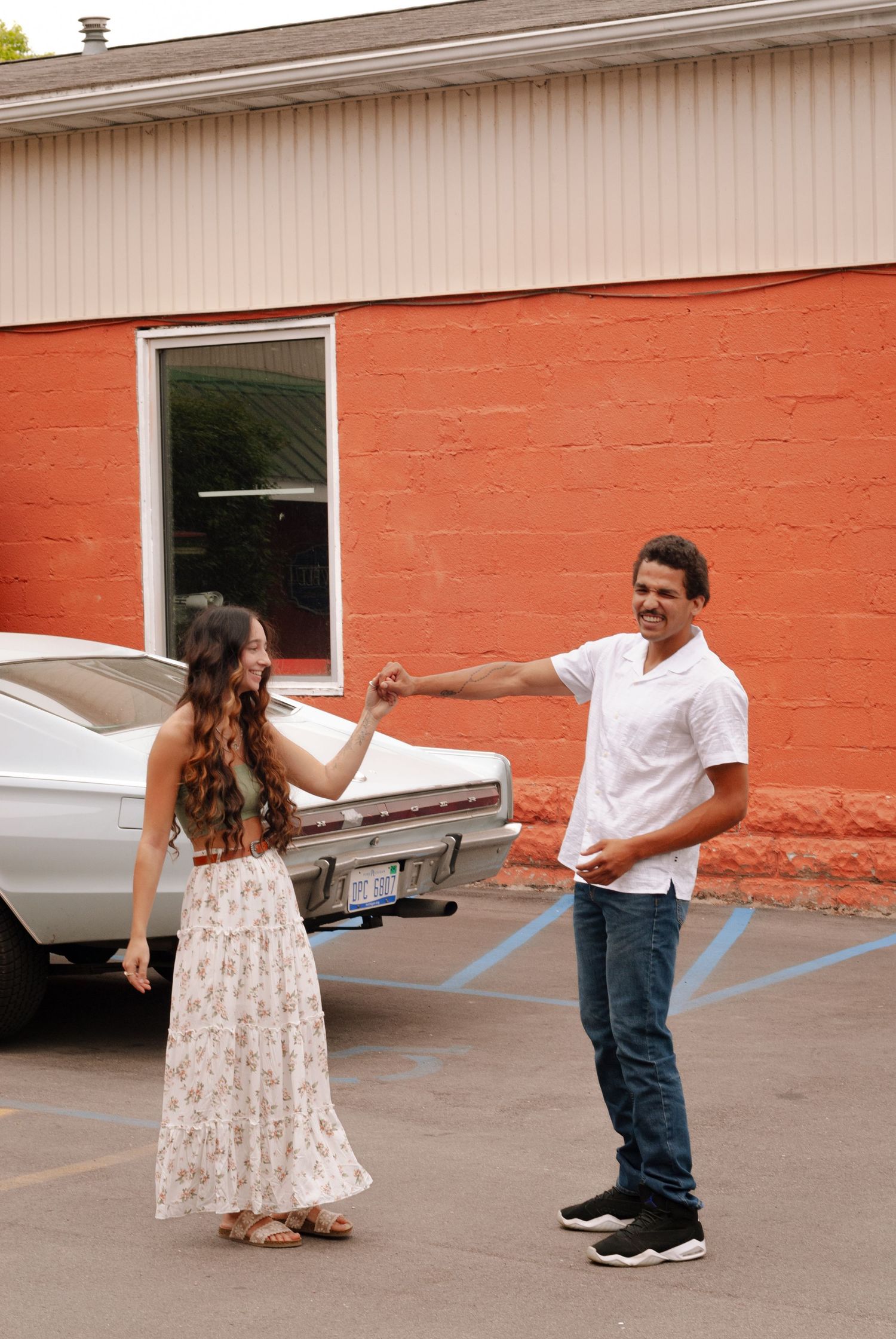 Smiling couple dancing and holding hands in a parking lot beside a vintage white car.