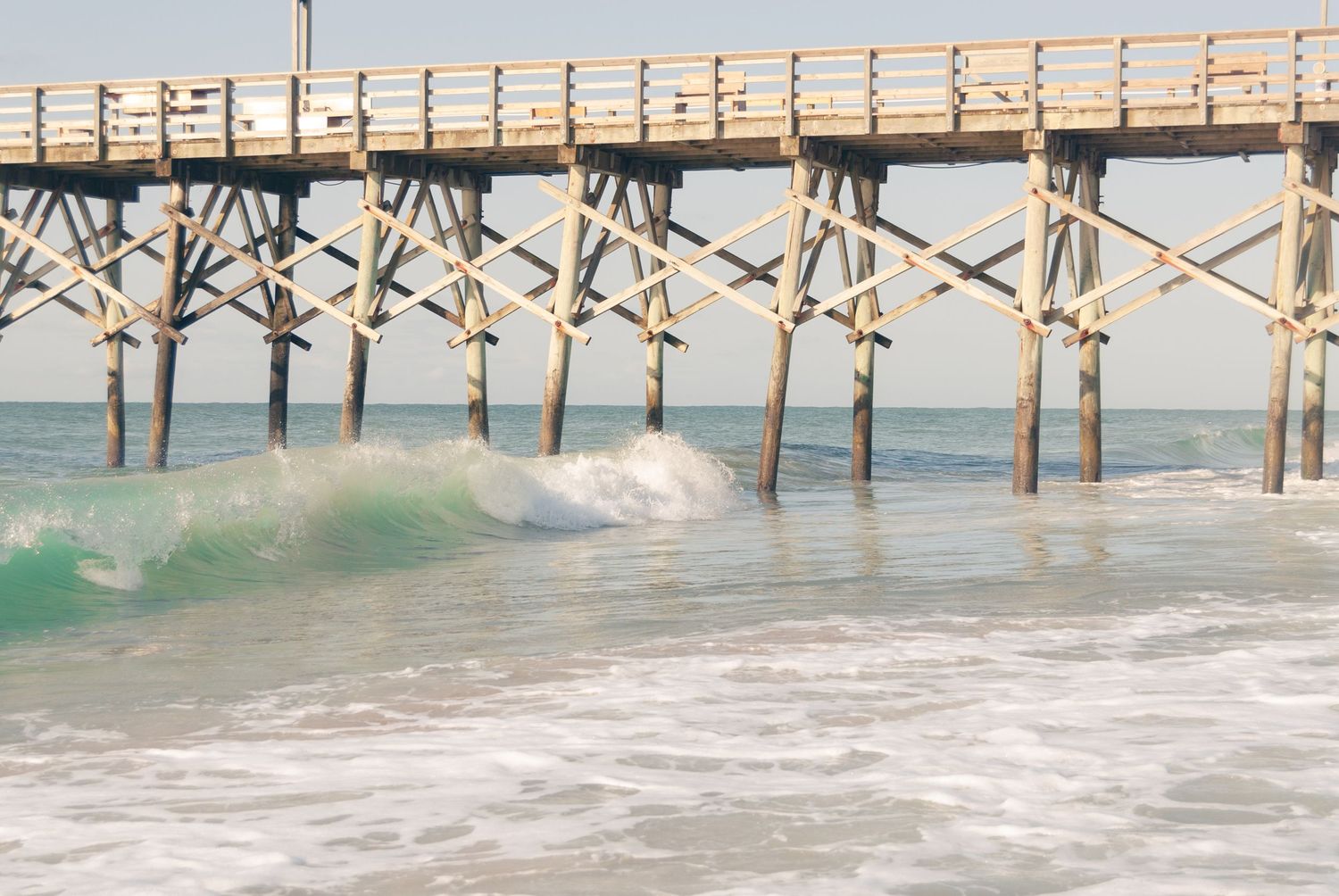 Wooden beach pier with crashing turquoise waves beneath its structure on a sunny day.