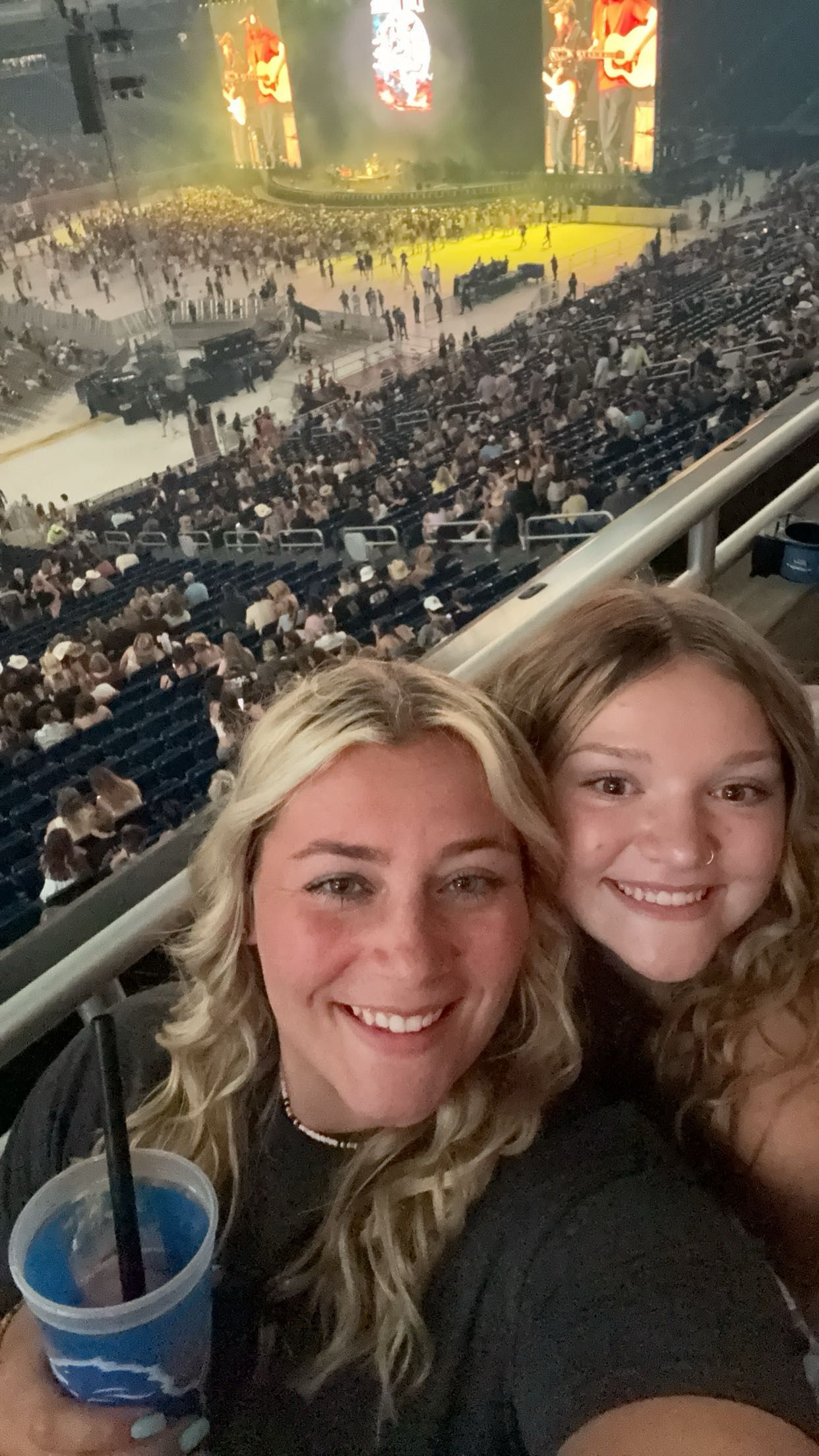 Two smiling women take a selfie at a packed indoor concert arena with a bright stage in the background.