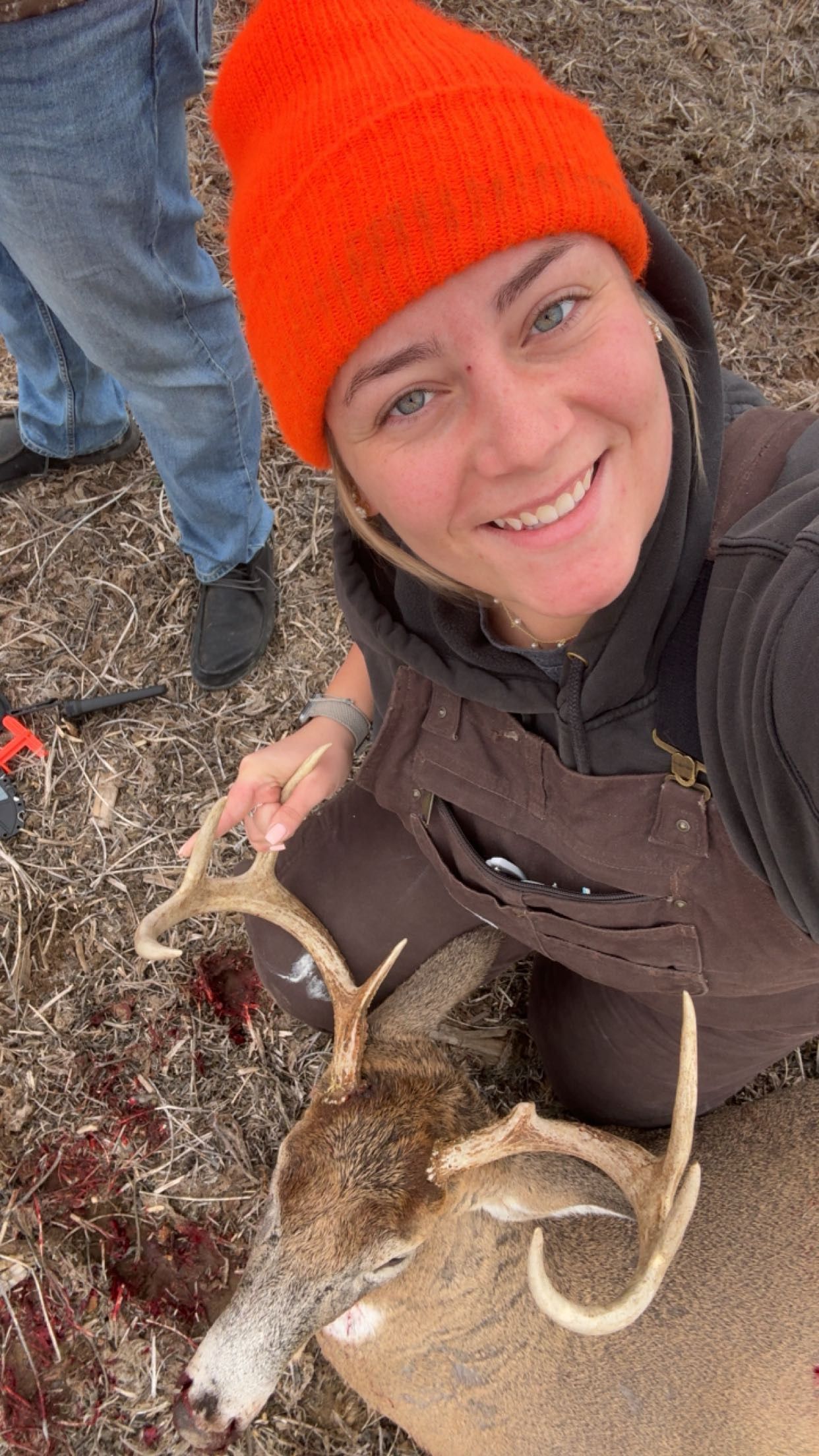 Woman in orange beanie smiles holding antlers of harvested whitetail buck deer during hunting season outdoors.