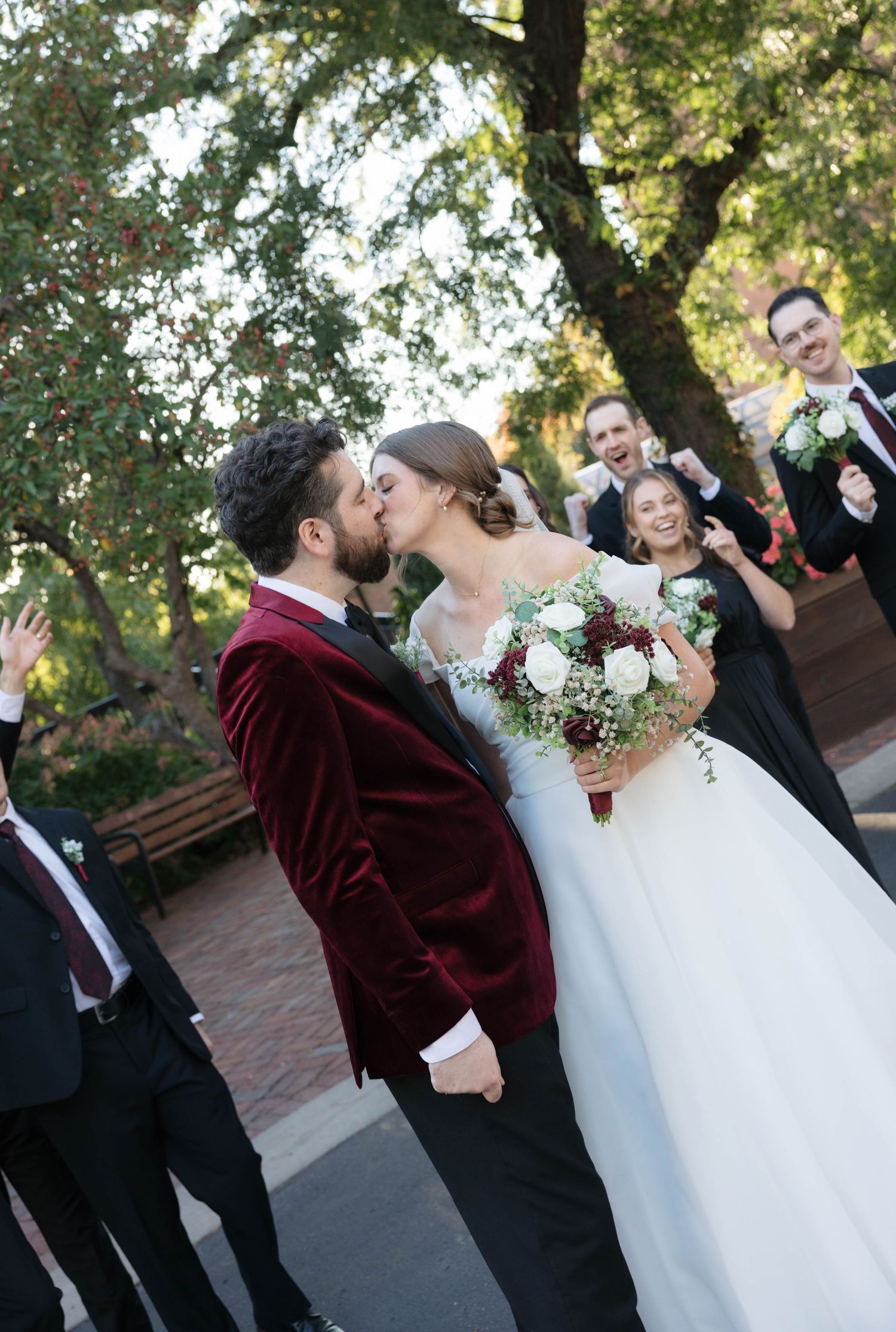 Groom in a burgundy velvet suit kisses his bride as the wedding party celebrates with confetti in an outdoor setting.
