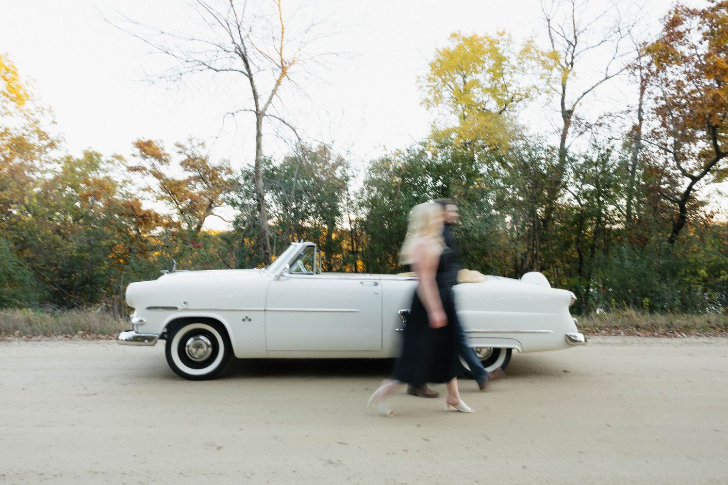 A blurred couple walks past a vintage white convertible car surrounded by autumn trees on a sandy path.