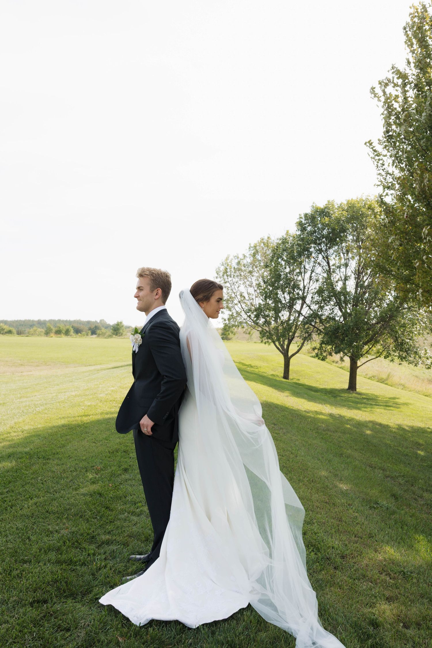 Bride and groom standing back-to-back in a vast green field with flowing cathedral veil.