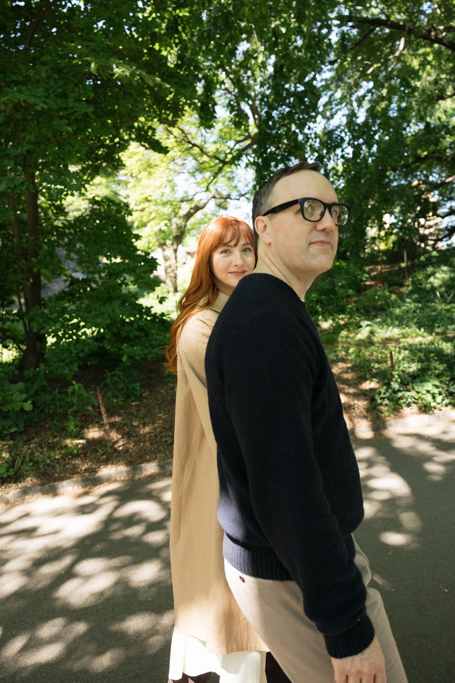 Couple standing back-to-back on a sunlit park path, surrounded by lush green trees and dappled shade.