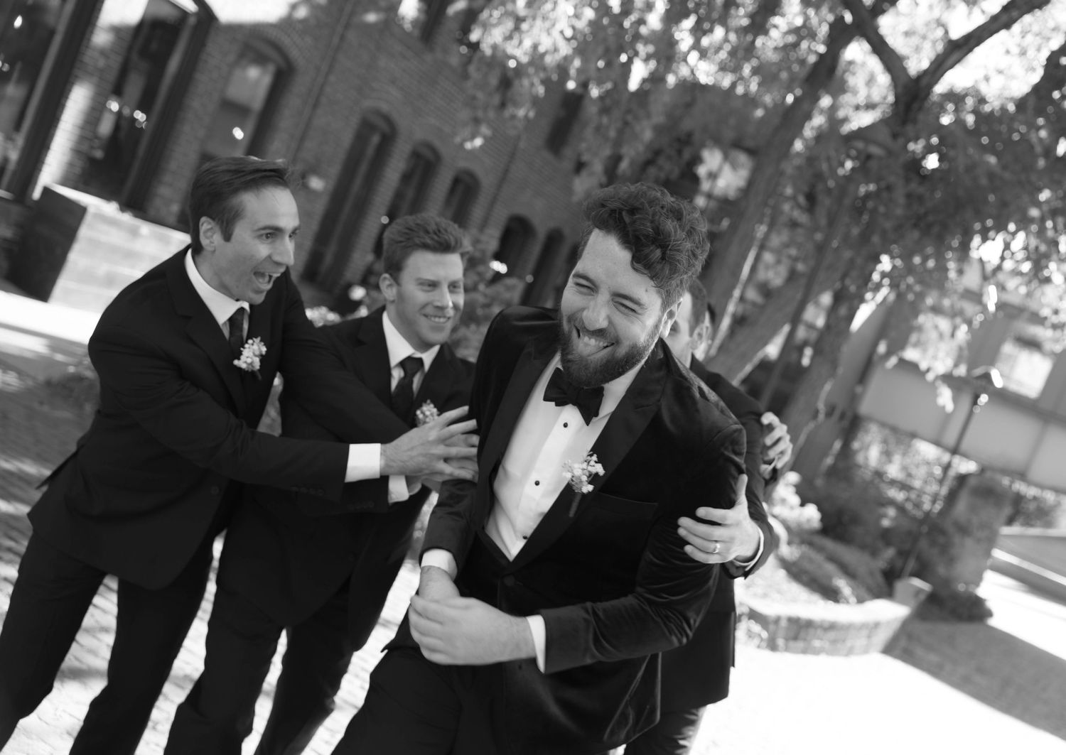 Black and white photo of groomsmen in tuxedos playfully celebrating with the groom outdoors before the wedding ceremony.