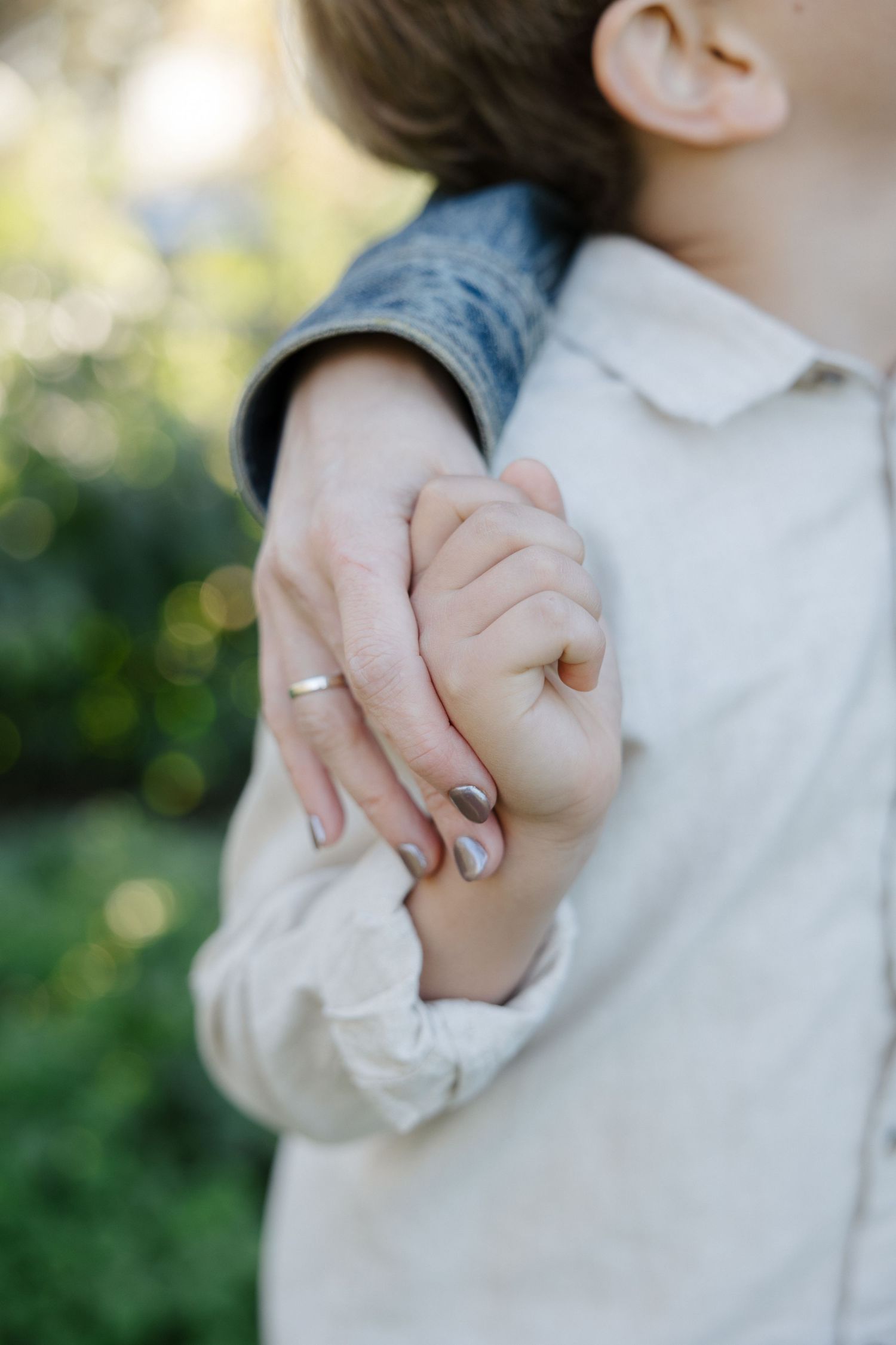 A child holds a woman's hand with a wedding ring in a lush green outdoor setting.