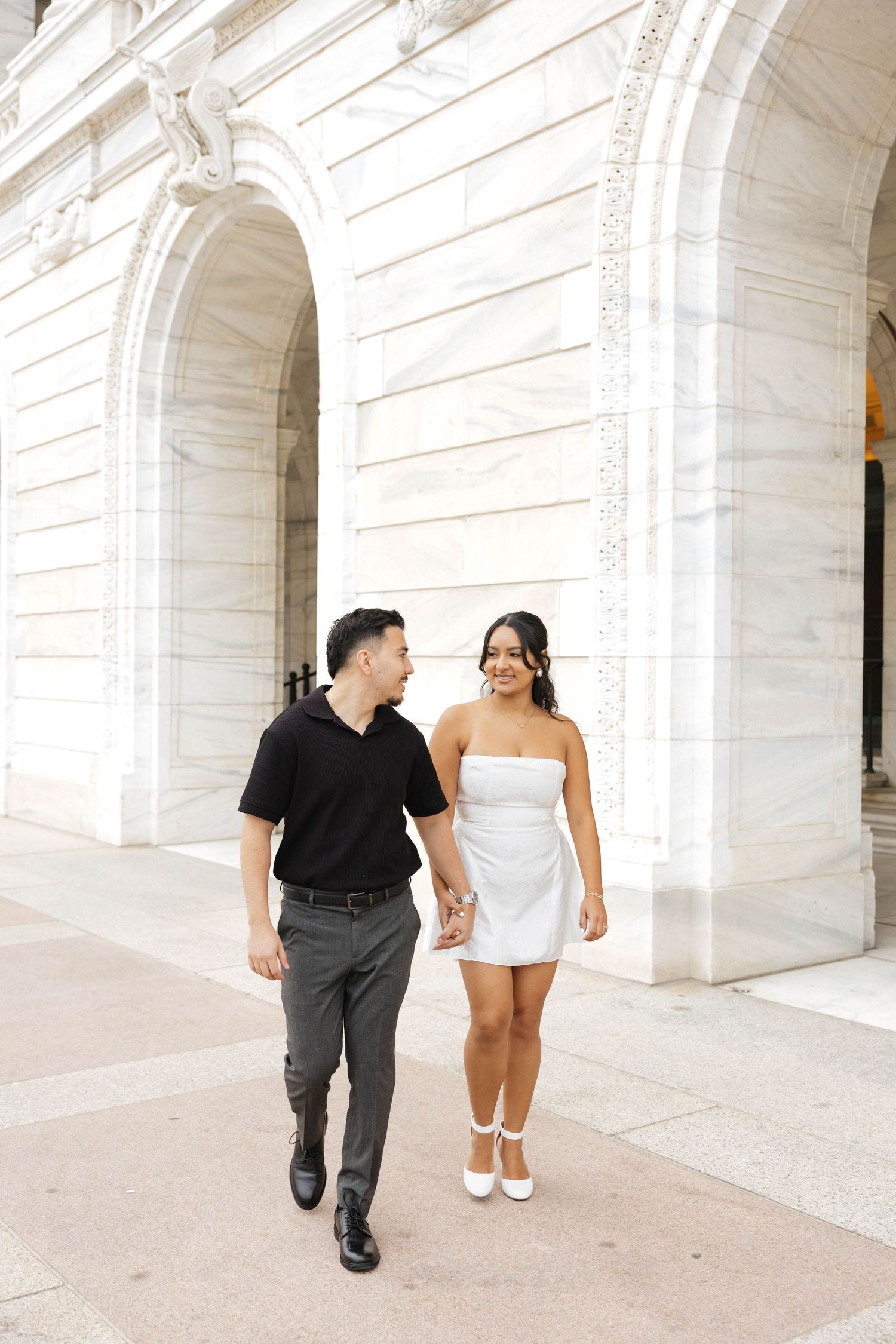 A couple walks hand in hand past white marble arches, man in black shirt and woman in white mini dress.