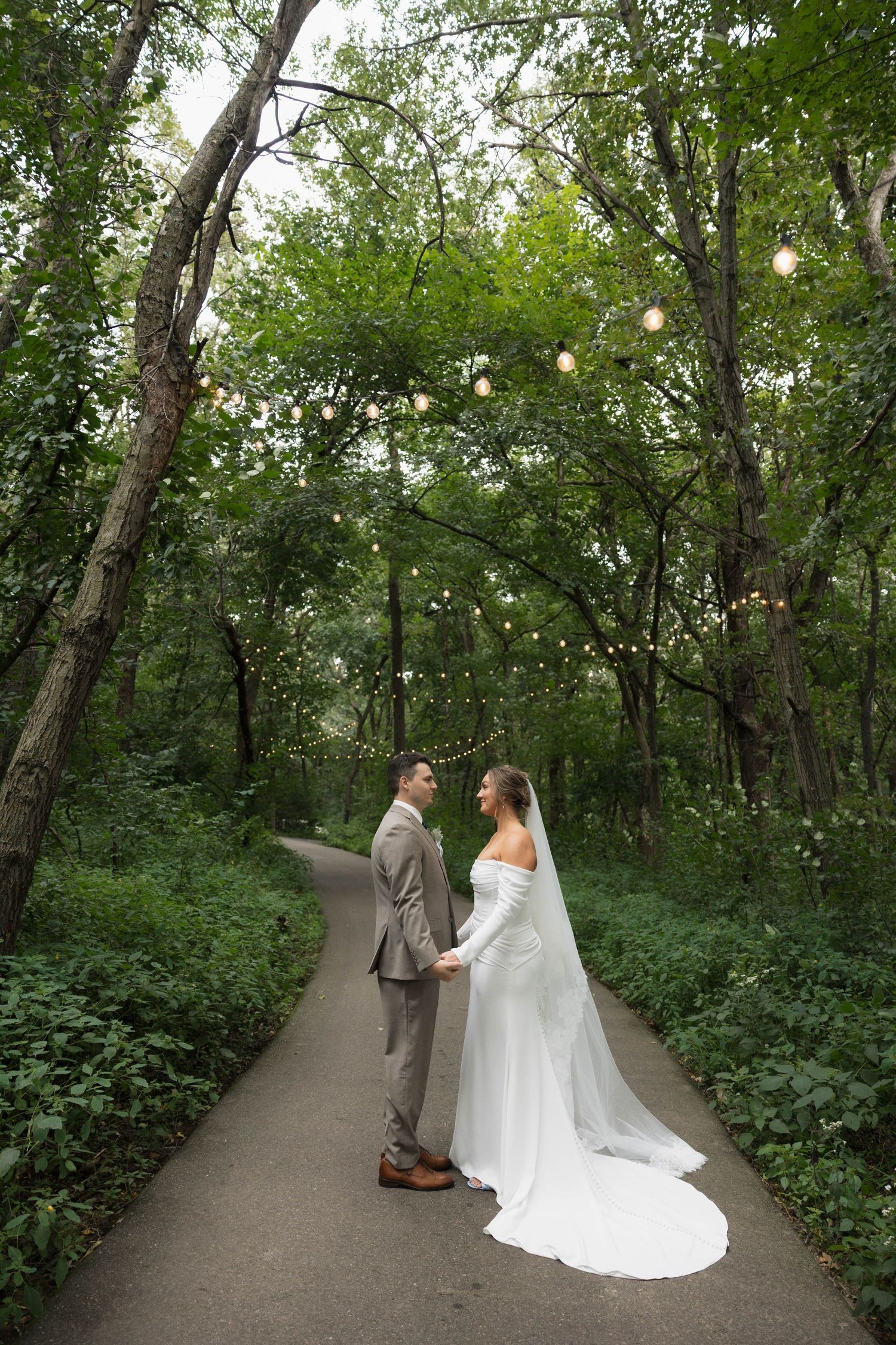 Bride and groom holding hands on a tree-lined path adorned with string lights at wedding.