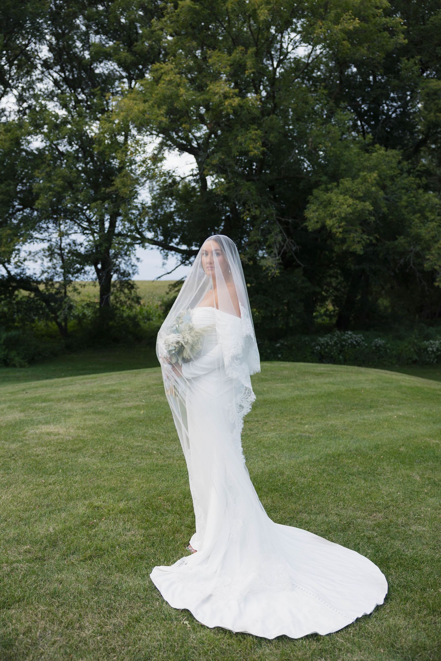 Bride in white fitted gown and long veil stands on green lawn surrounded by lush trees.