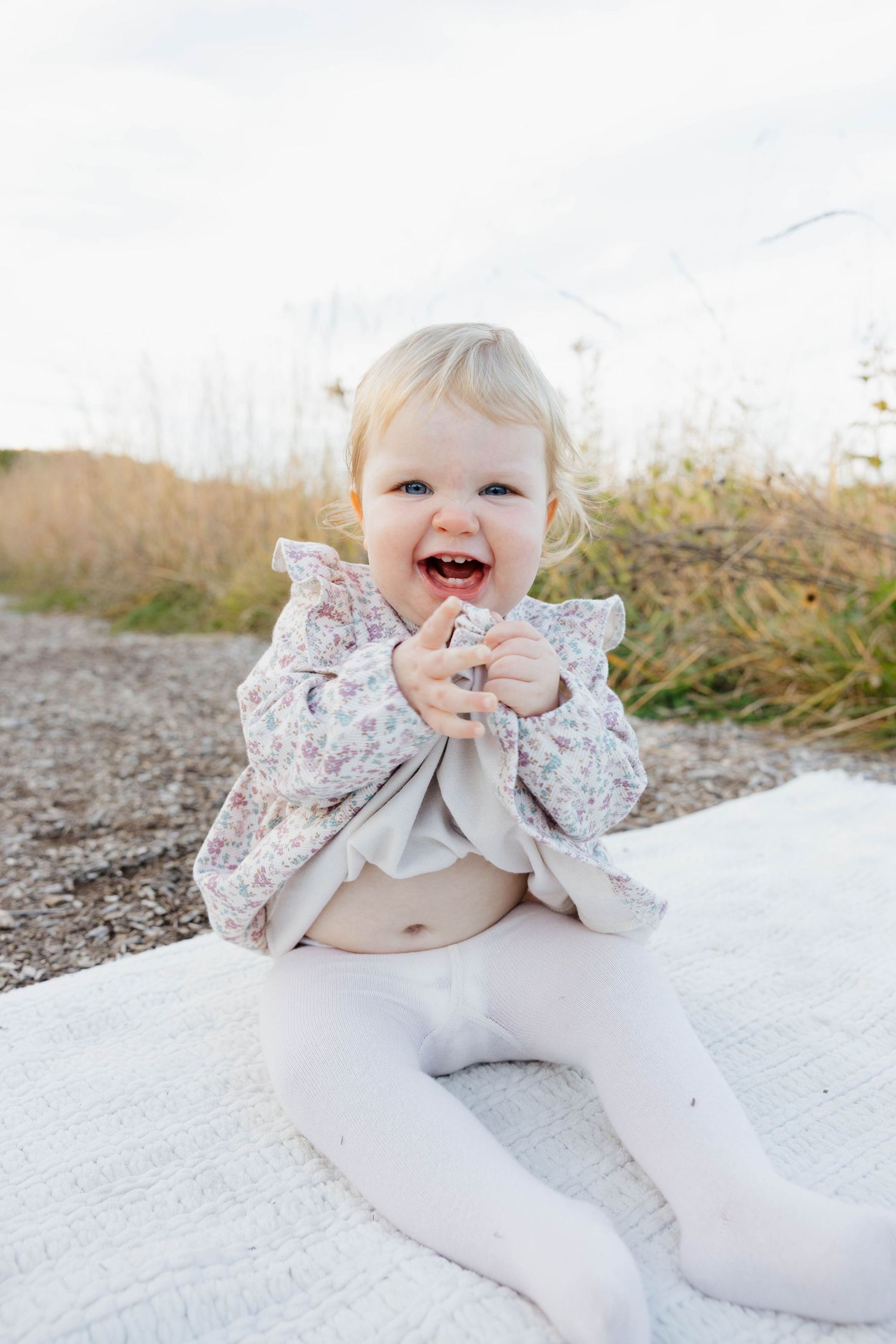 A laughing blonde baby girl sits on a white blanket outdoors in a floral top.