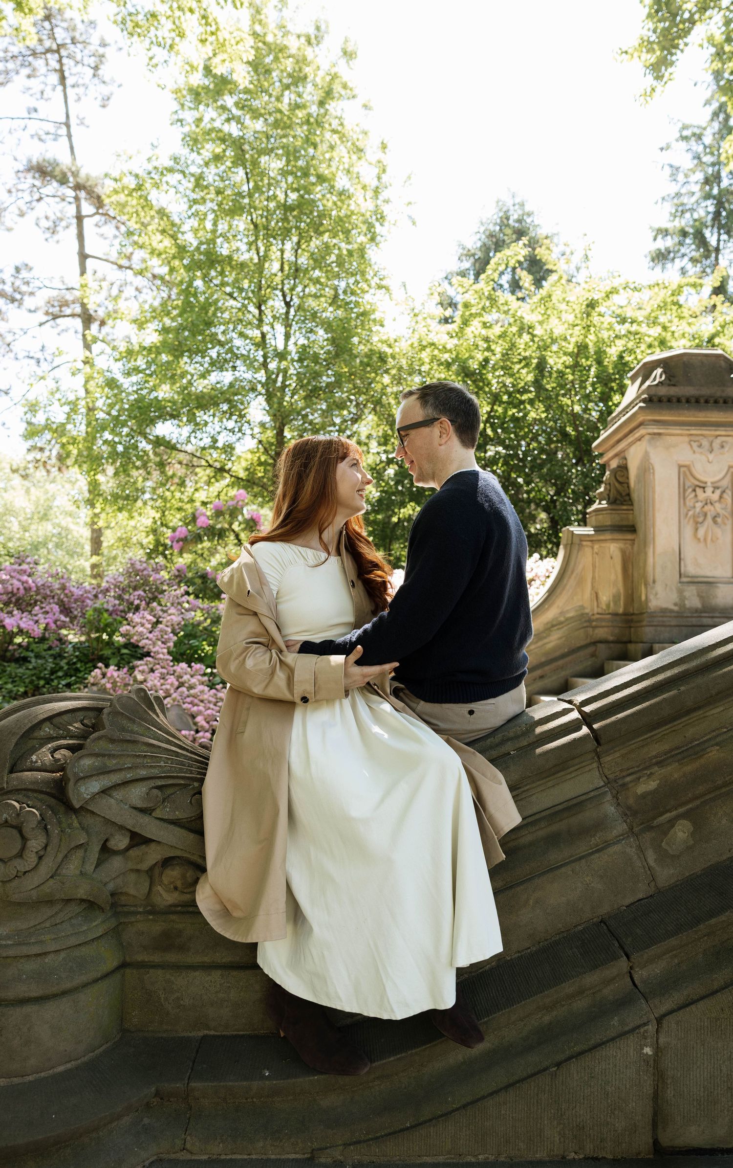 Couple sitting on ornate stone steps in a park, surrounded by blooming flowers and lush green trees.