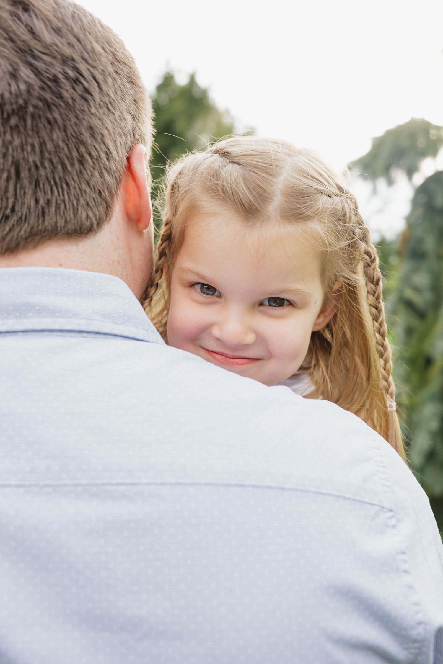 A smiling little girl with braided hair rests her head on her father's shoulder outdoors.