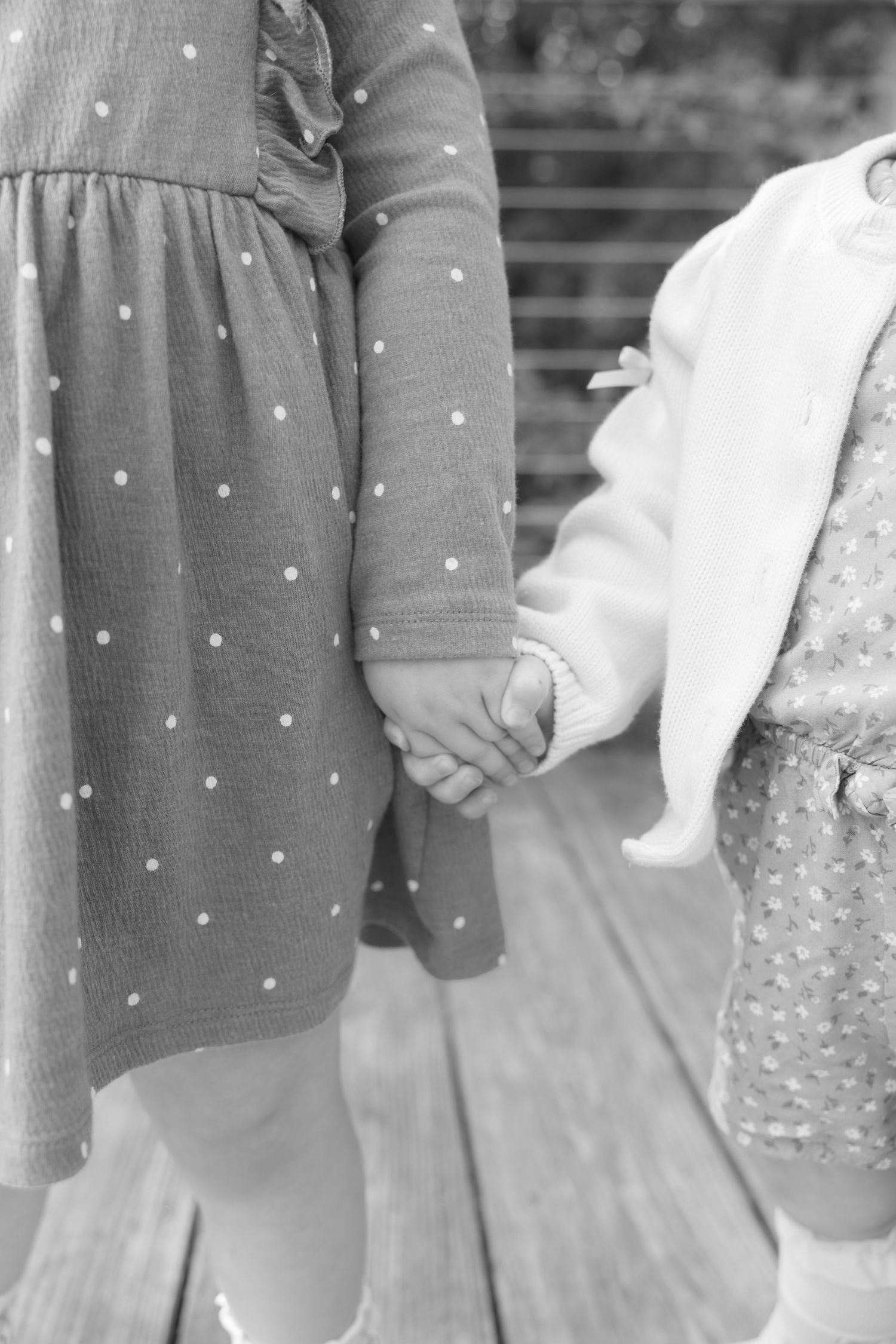 Black and white photo of two young girls in polka dot dresses holding hands on a wooden deck.