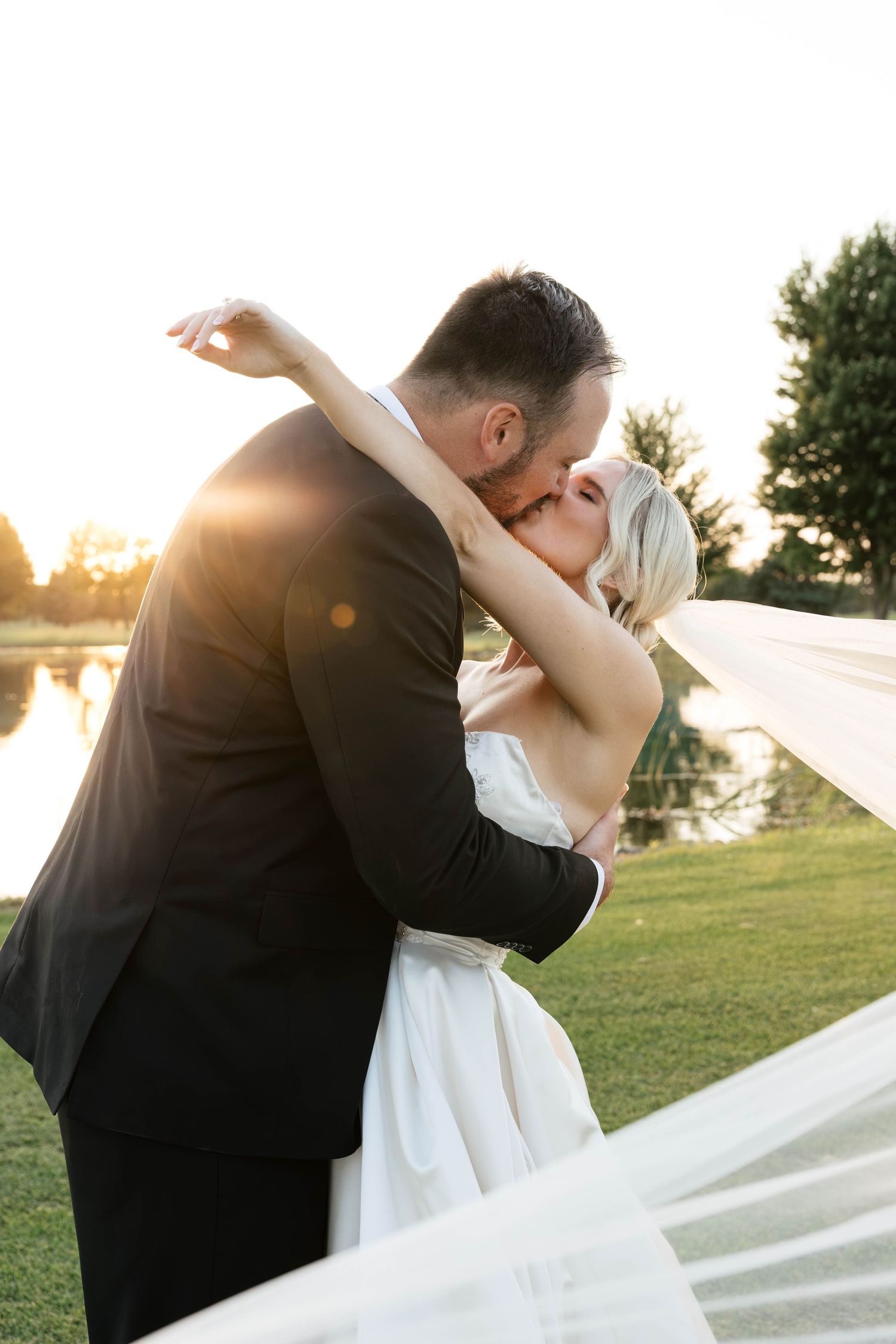 Bride and groom sharing a romantic kiss at golden sunset by a serene pond, veil flowing in the breeze.