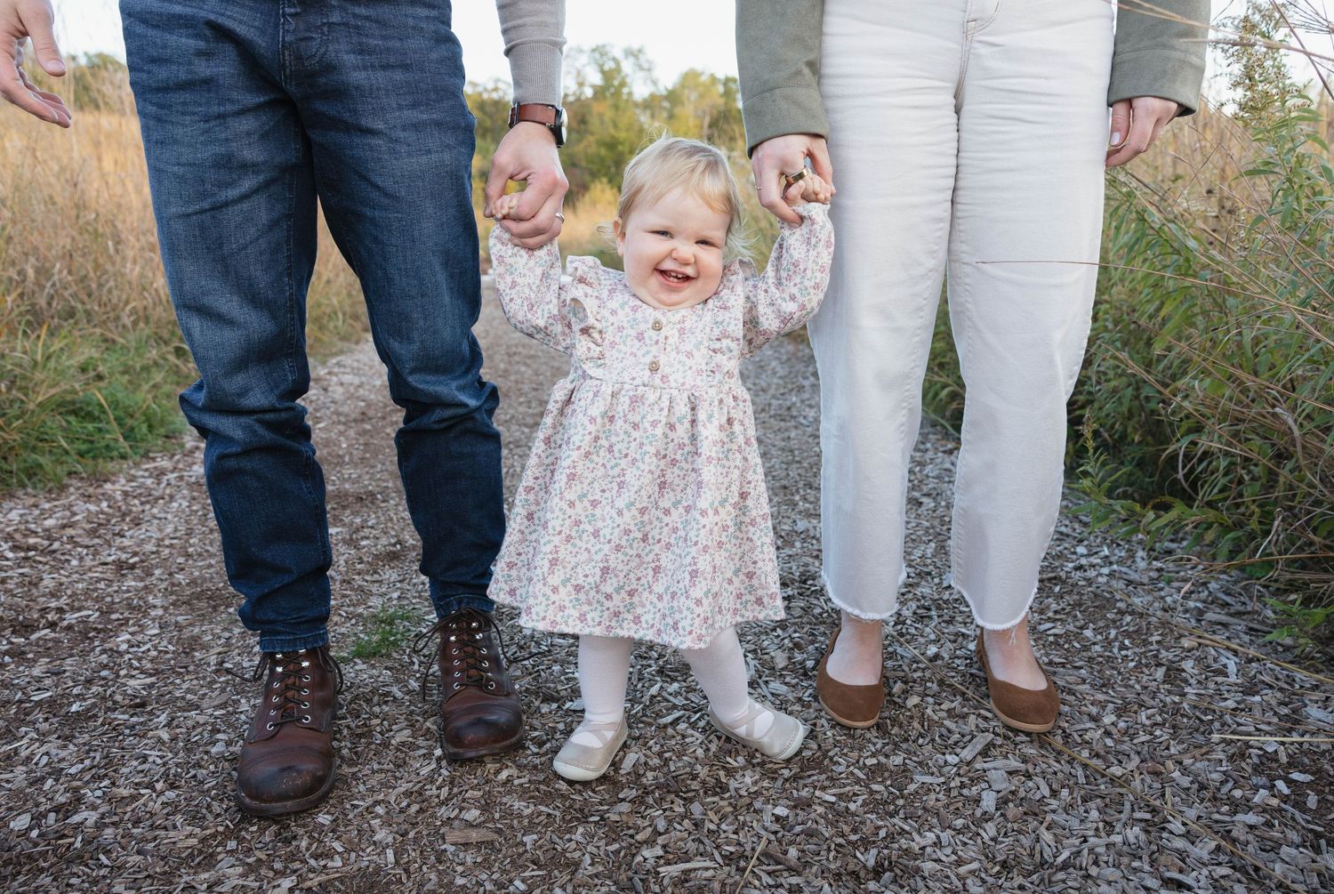 Proud parents hold their laughing toddler girl's hands as she stands on a gravel path outdoors.