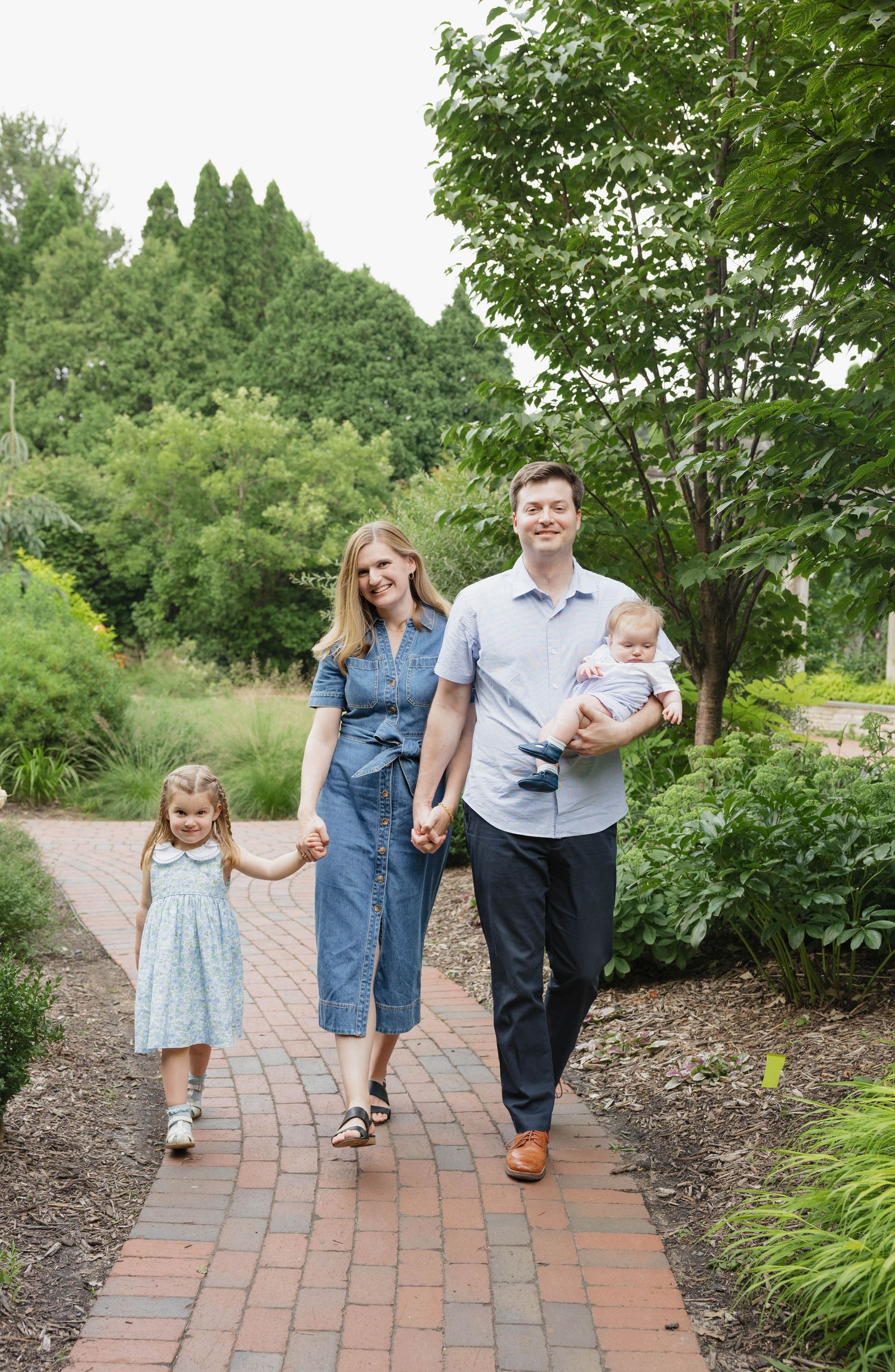 A happy family of four walks hand-in-hand along a brick garden path surrounded by lush greenery.