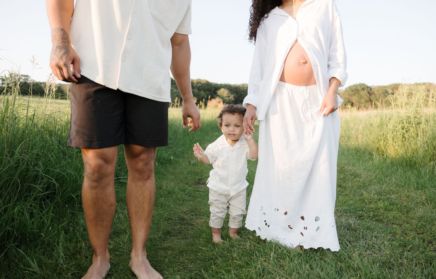 A pregnant woman in white and a toddler stand with a man in a green field.