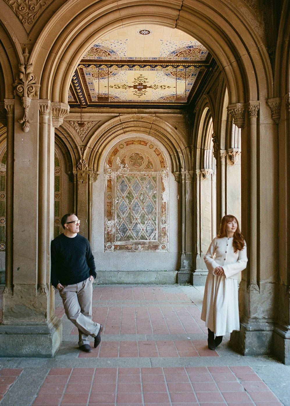 Couple posing apart under ornate arched arcade with colorful mosaic ceiling at Bethesda Terrace, Central Park.