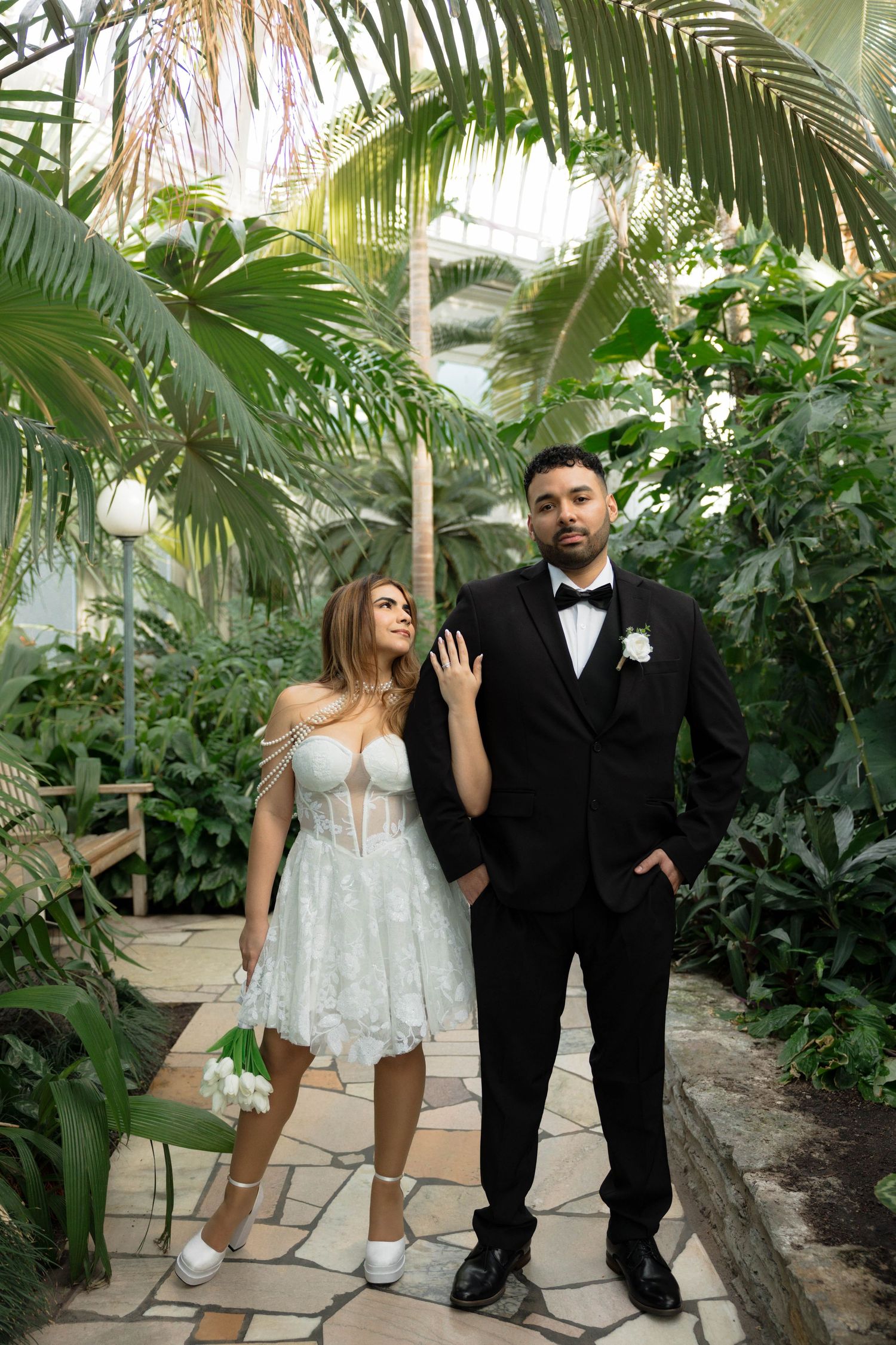 Bride in short lace dress and groom in black tuxedo pose in tropical greenhouse.