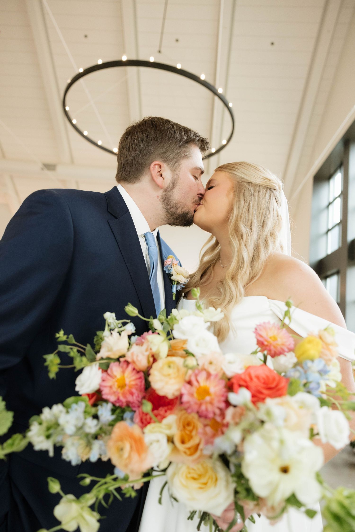 Bride and groom share a kiss holding a vibrant colorful floral bouquet indoors.