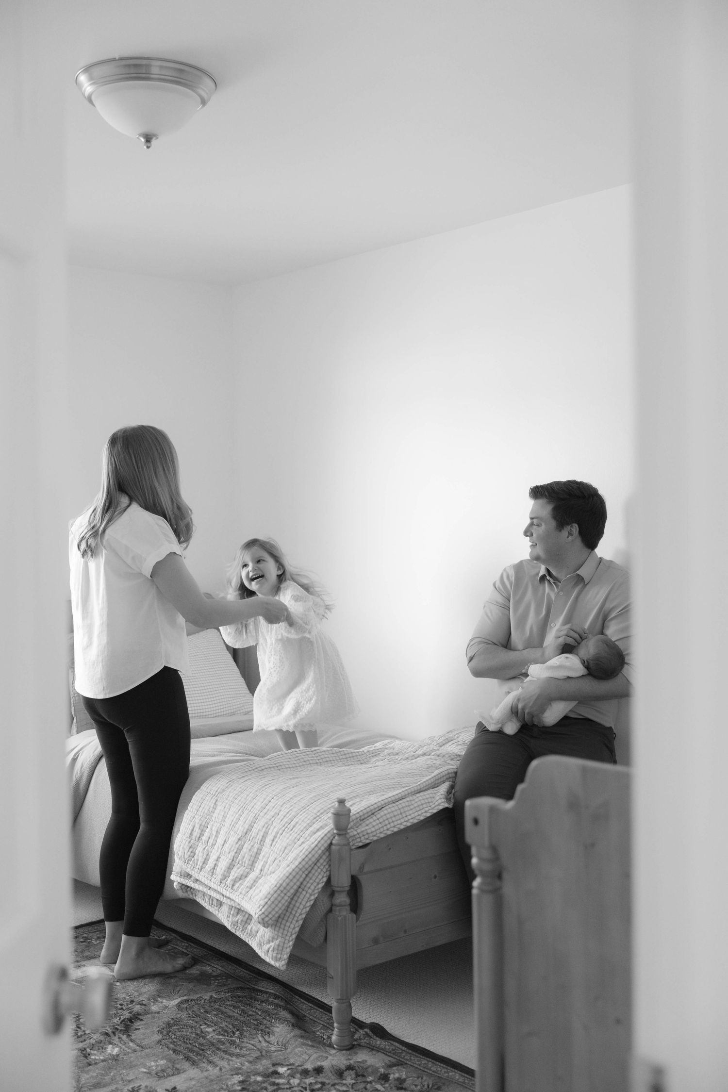 Black and white family photo of parents playing with young children on bed in cozy bedroom interior.