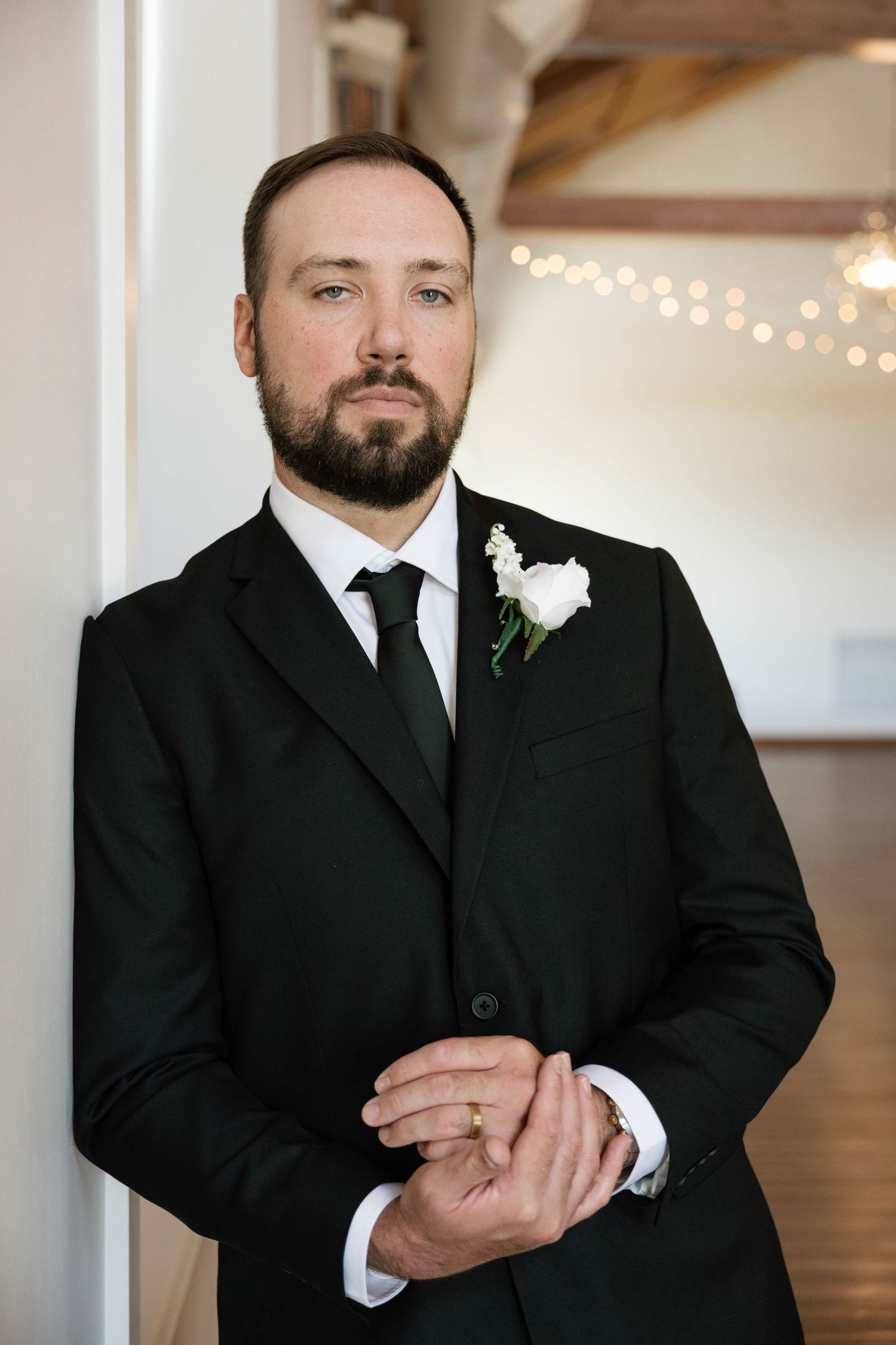 Dapper groom in black suit with white boutonniere leaning against a column in an elegant wedding venue.