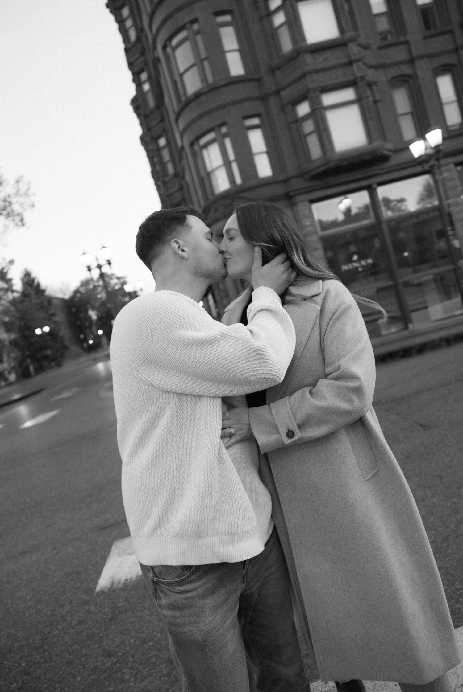 Black and white photo of a couple sharing a kiss on a city street at dusk near a historic building.