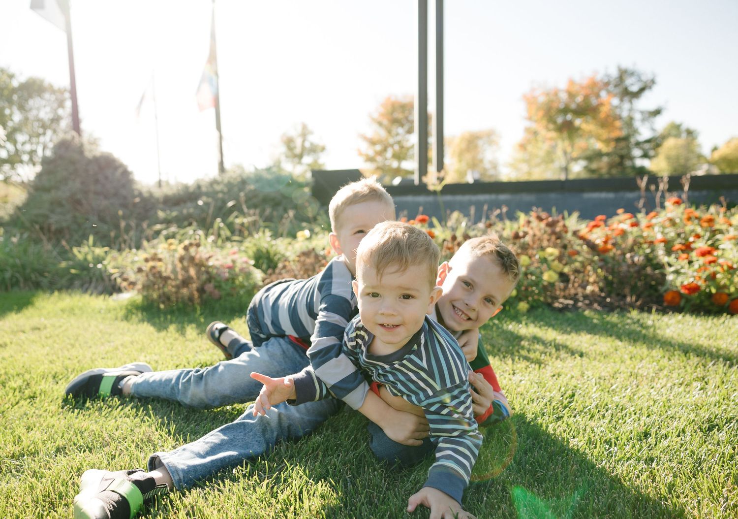 Three young boys in matching striped outfits playfully pile on each other in a sunny garden.