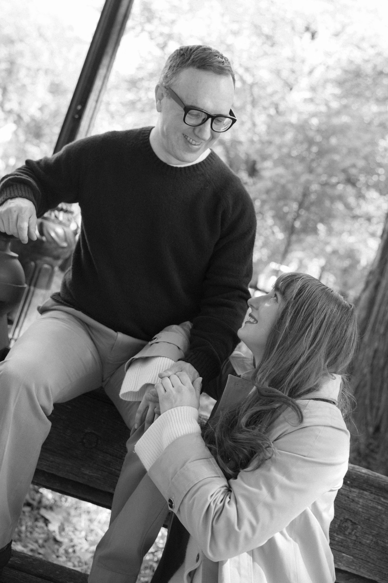 Black and white photo of smiling couple sharing a joyful moment on a wooden swing in the park.