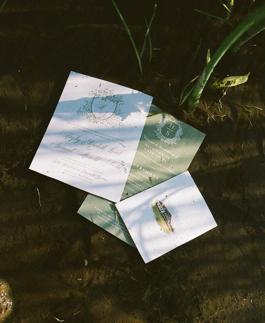 Elegant watercolor wedding invitations and a gold engagement ring rest on a natural stone surface surrounded by greenery.
