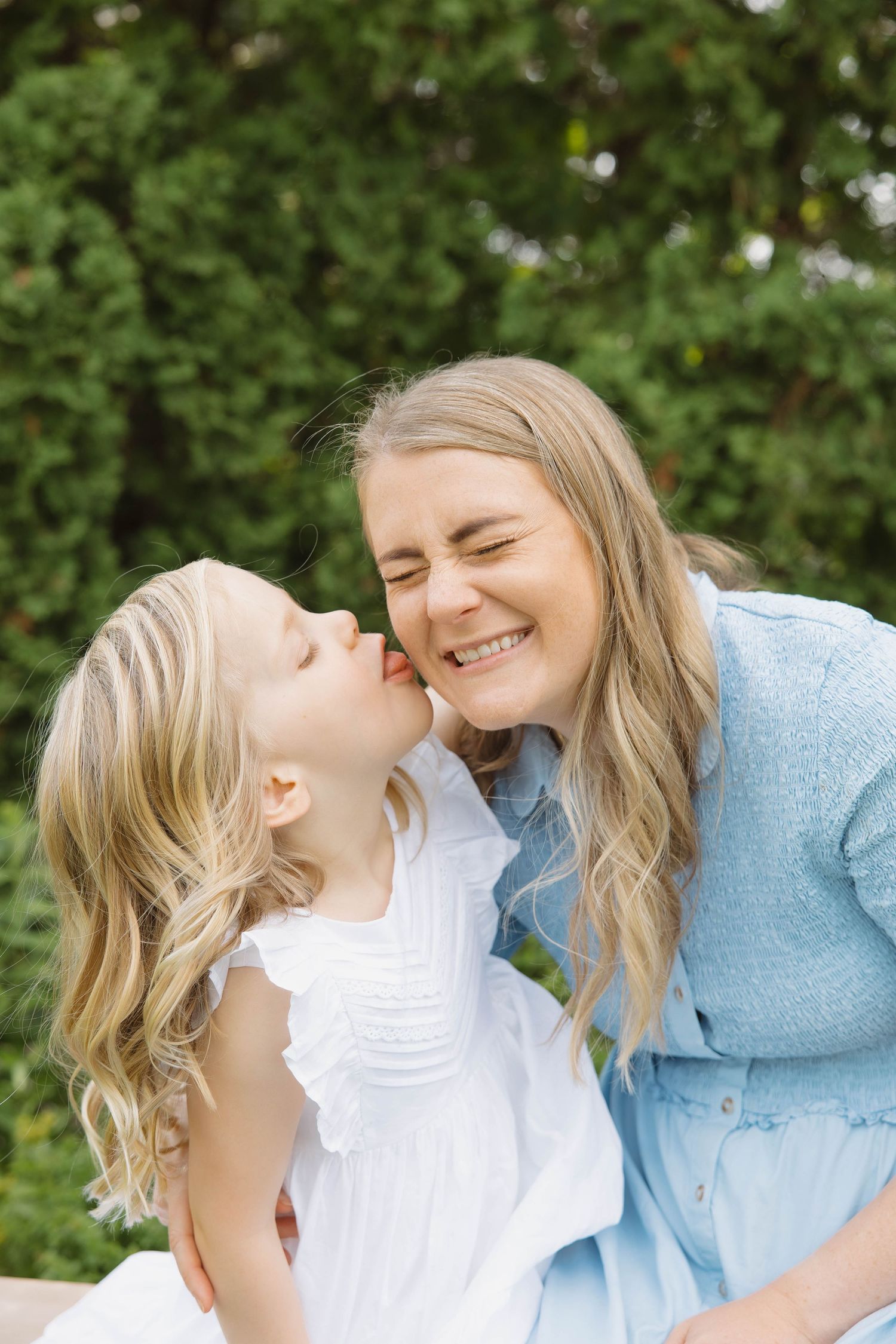 Little girl kissing her laughing mother on the cheek outdoors near green trees.