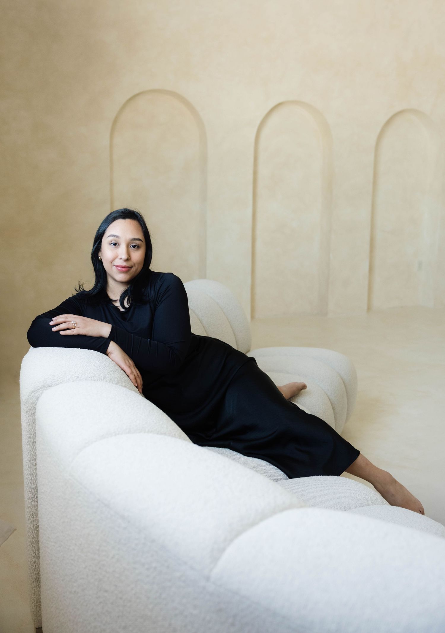 Woman in black dress reclines on white boucle sofa against beige arched wall backdrop.